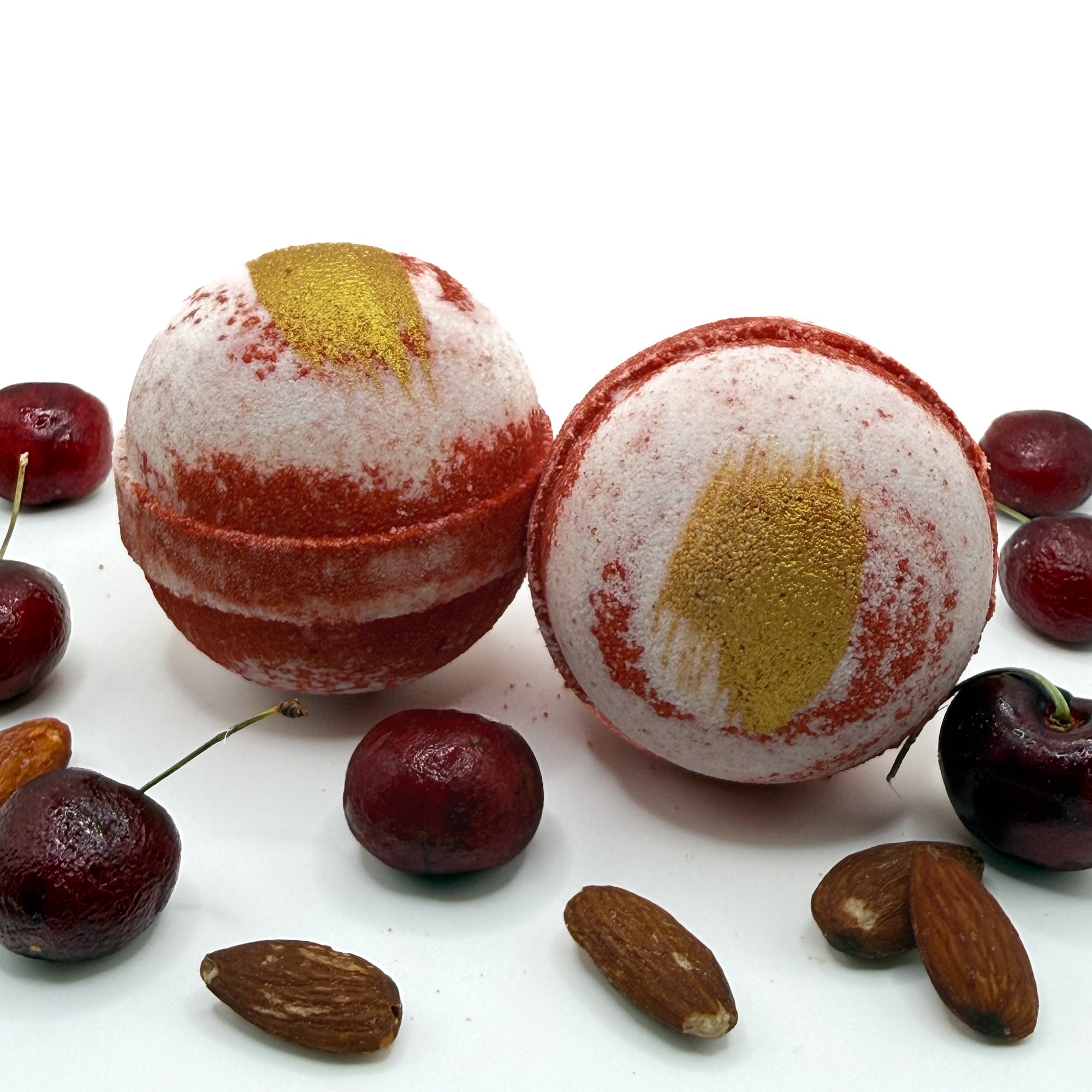 Two handmade cherry almond bath bombs with gold accents on a white background with cherries and almonds by Rusted Acre Soap Co.