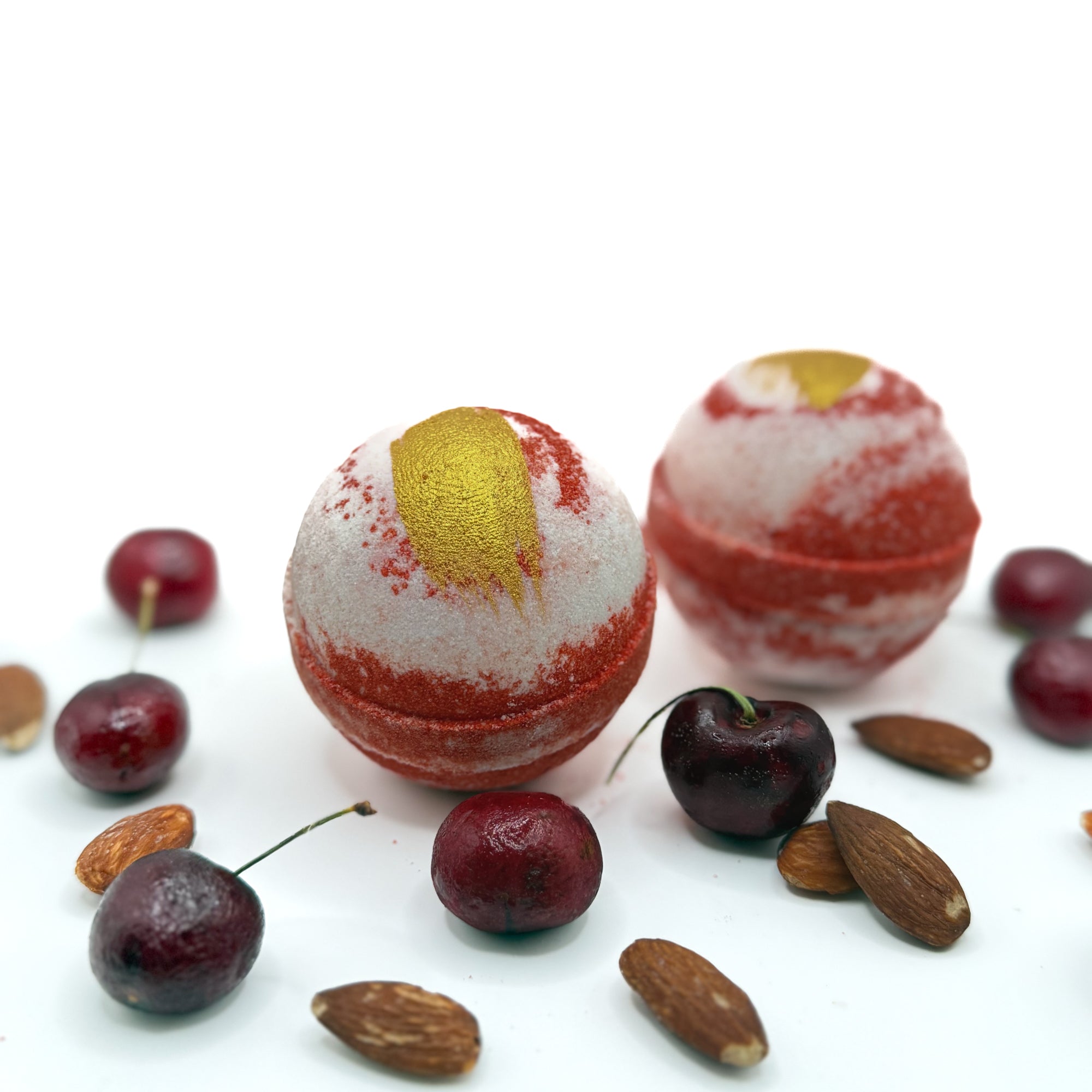 Two handmade cherry almond bath bombs with gold mica paint, surrounded by red cherries and almonds on a white background
