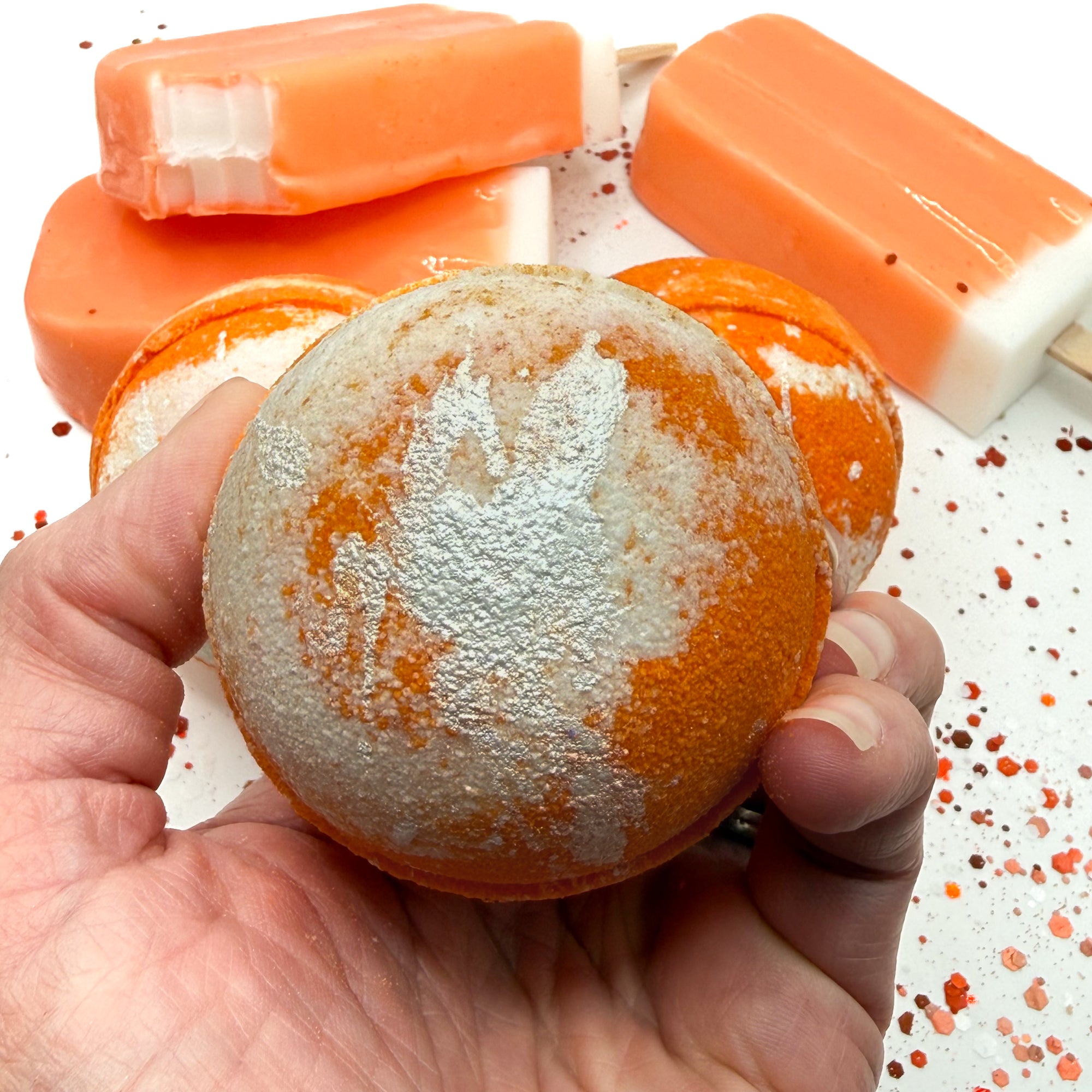 Orange and white bath bomb hand made by Rusted Acre Soap Co held in a hand with orange ice cream in the background.