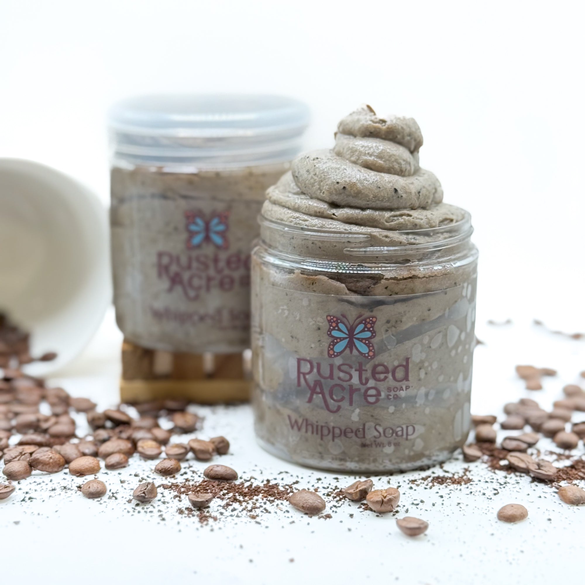 Two jars of Rusted Acre coffee whipped soap surrounded with coffee beans on a white background.