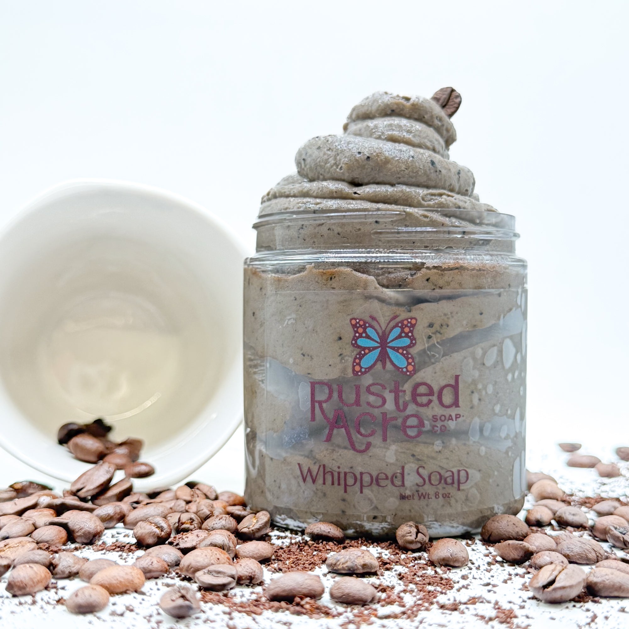 Jar of coffee whipped soap with 'Rusted Acre' branding, surrounded by coffee beans and a white mug on a light background.