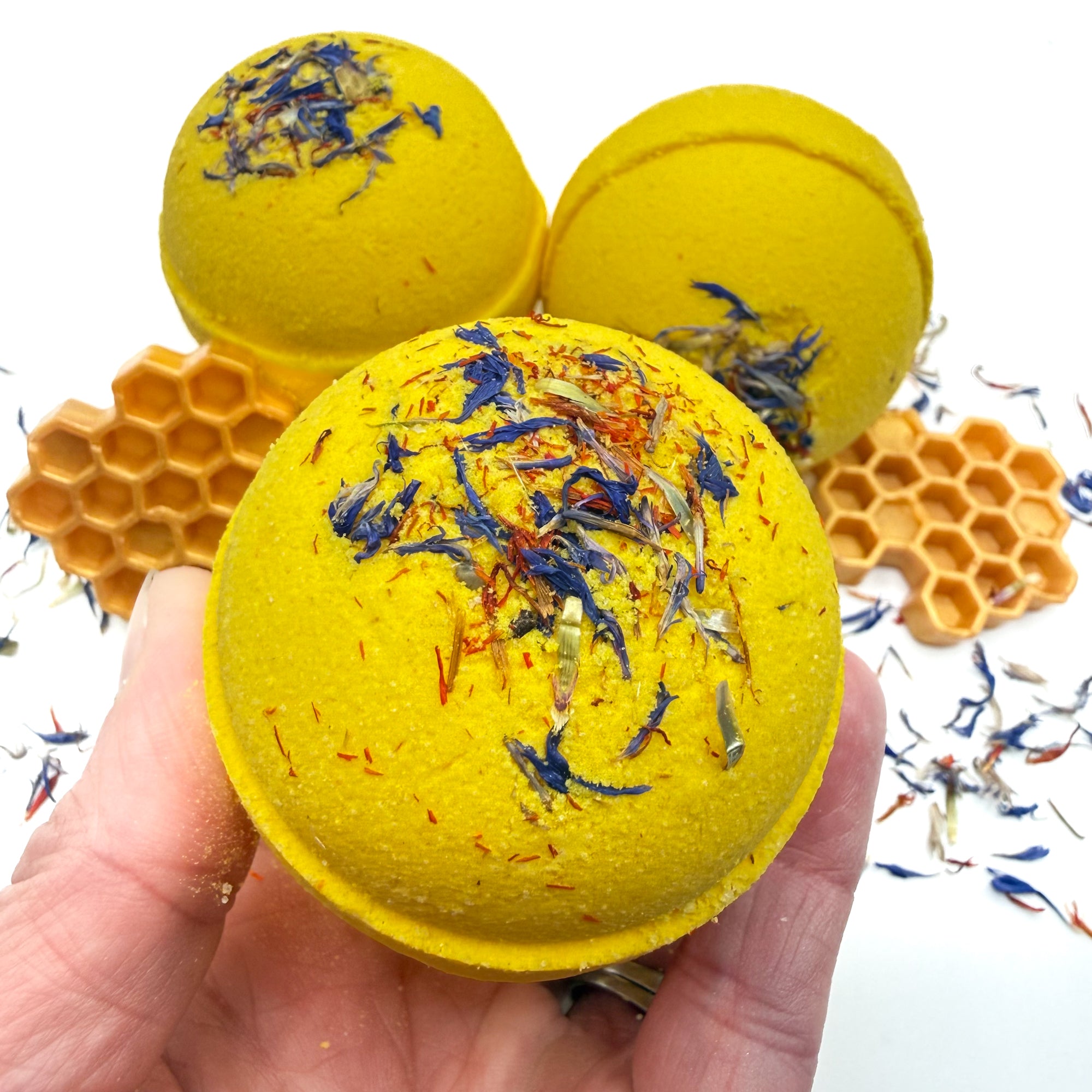 Yellow wildflower and honey bath bomb with embedded Wild flowers being held in a hand over a white background with wildflowers.