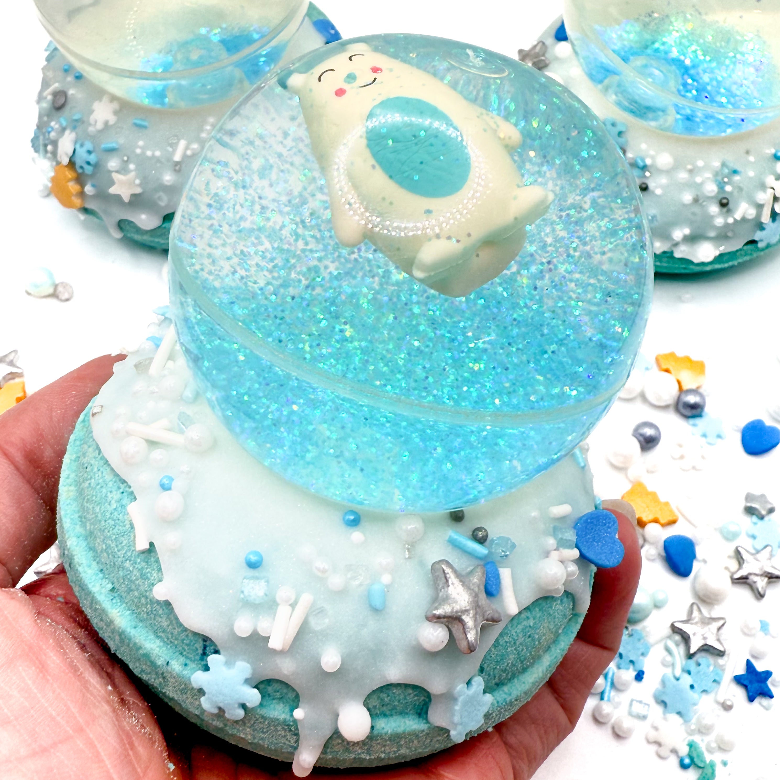 Snow globe bath bomb topped with an Abominable Snowman bouncy ball in a hand. Blue and silver sprinkles in the background.