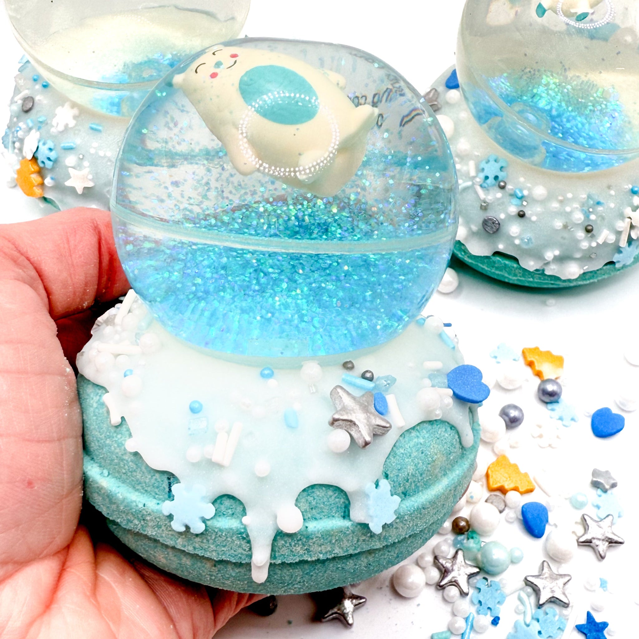 Snow globe bath bomb topped with an Abominable Snowman bouncy ball in a hand. Blue and silver sprinkles in the background.