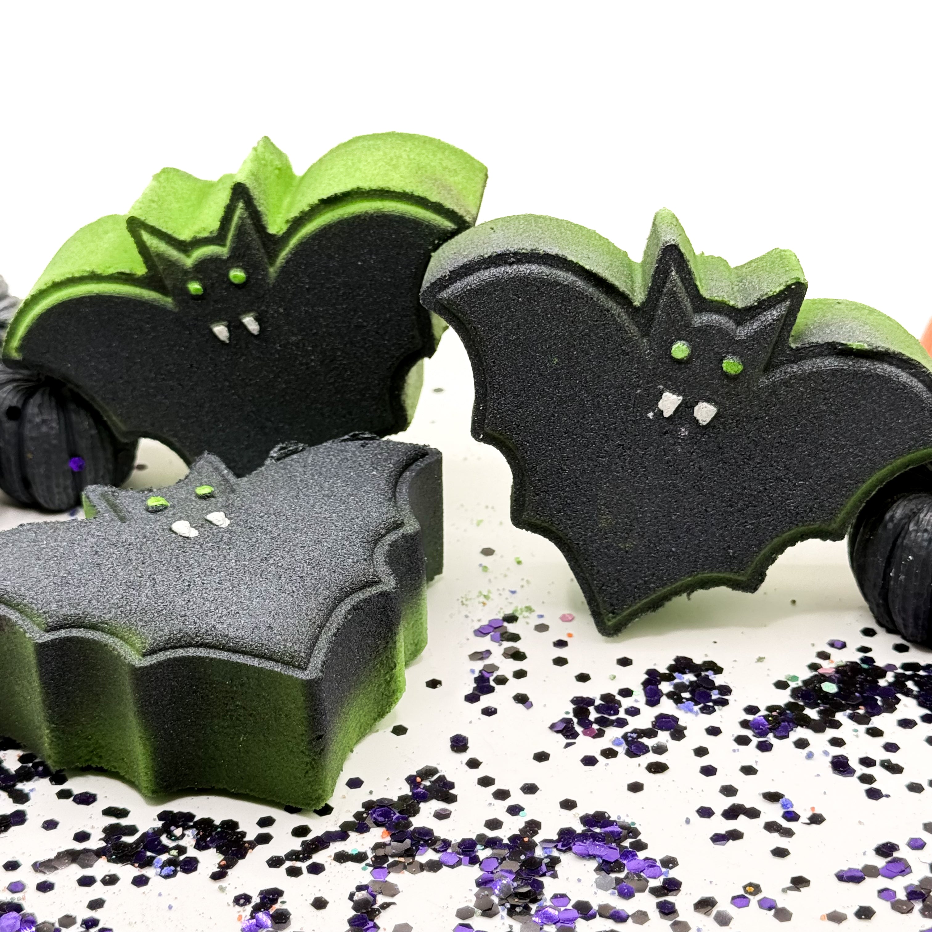Three bat bath bombs with black and green, decorated with small white fangs and green eyes, arranged on a white surface with scattered black and purple glitter.