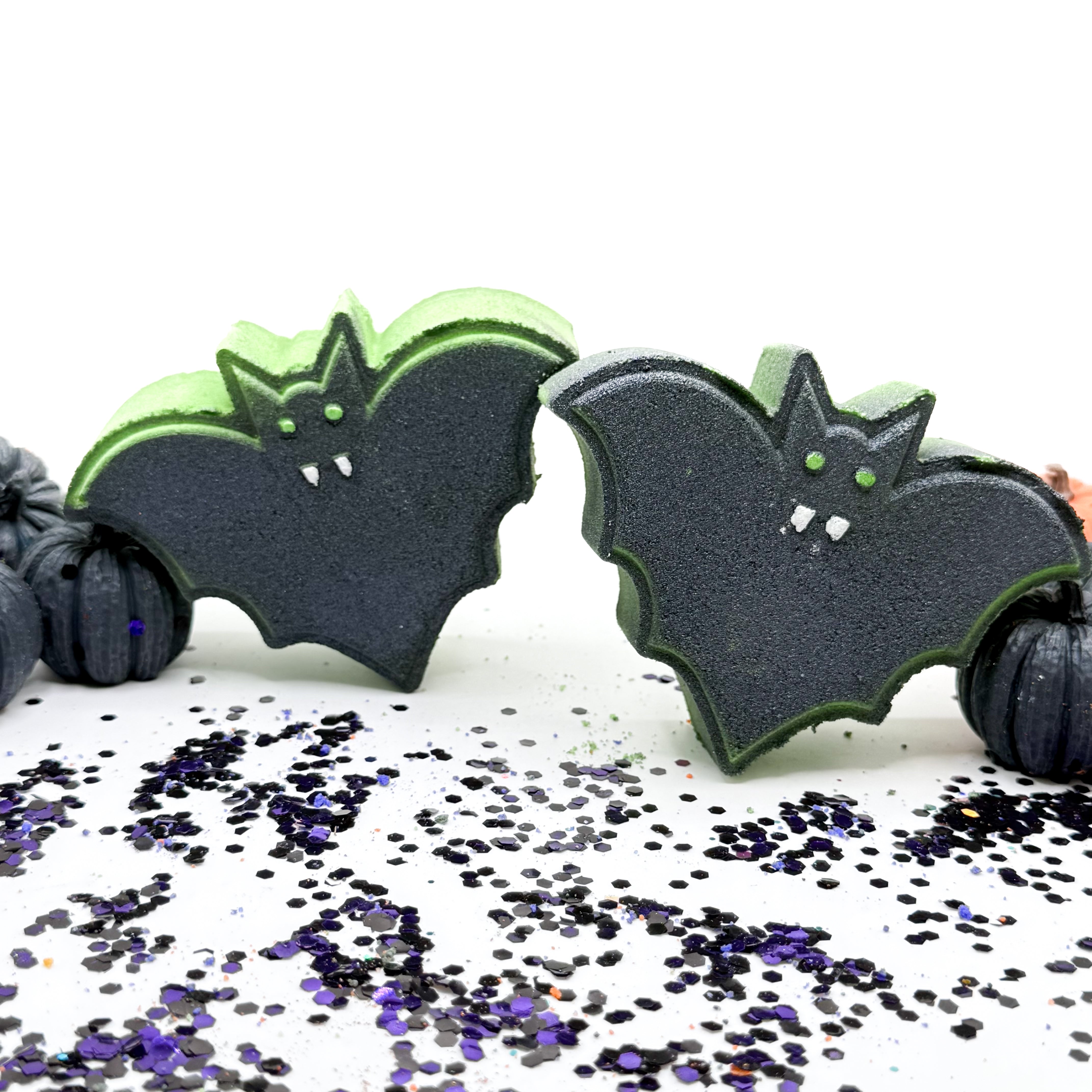 Two bat bath bombs with a black body and bright lime green edges, decorated with small white fangs and green eyes, displayed on a white surface scattered with black and purple glitter.