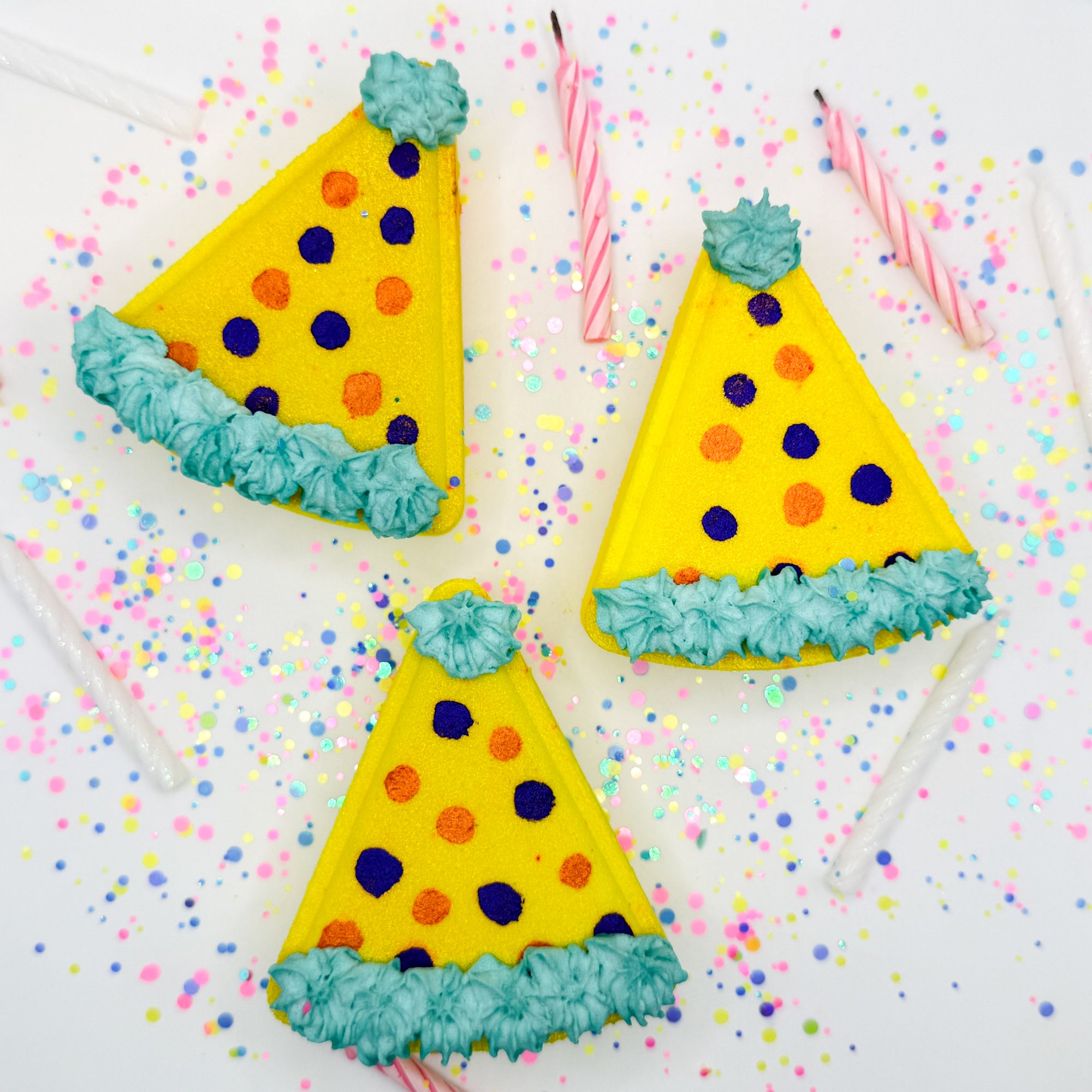 Three yellow birthday hat shaped bath bombs with hand painted polka dots lying flat on a white background.