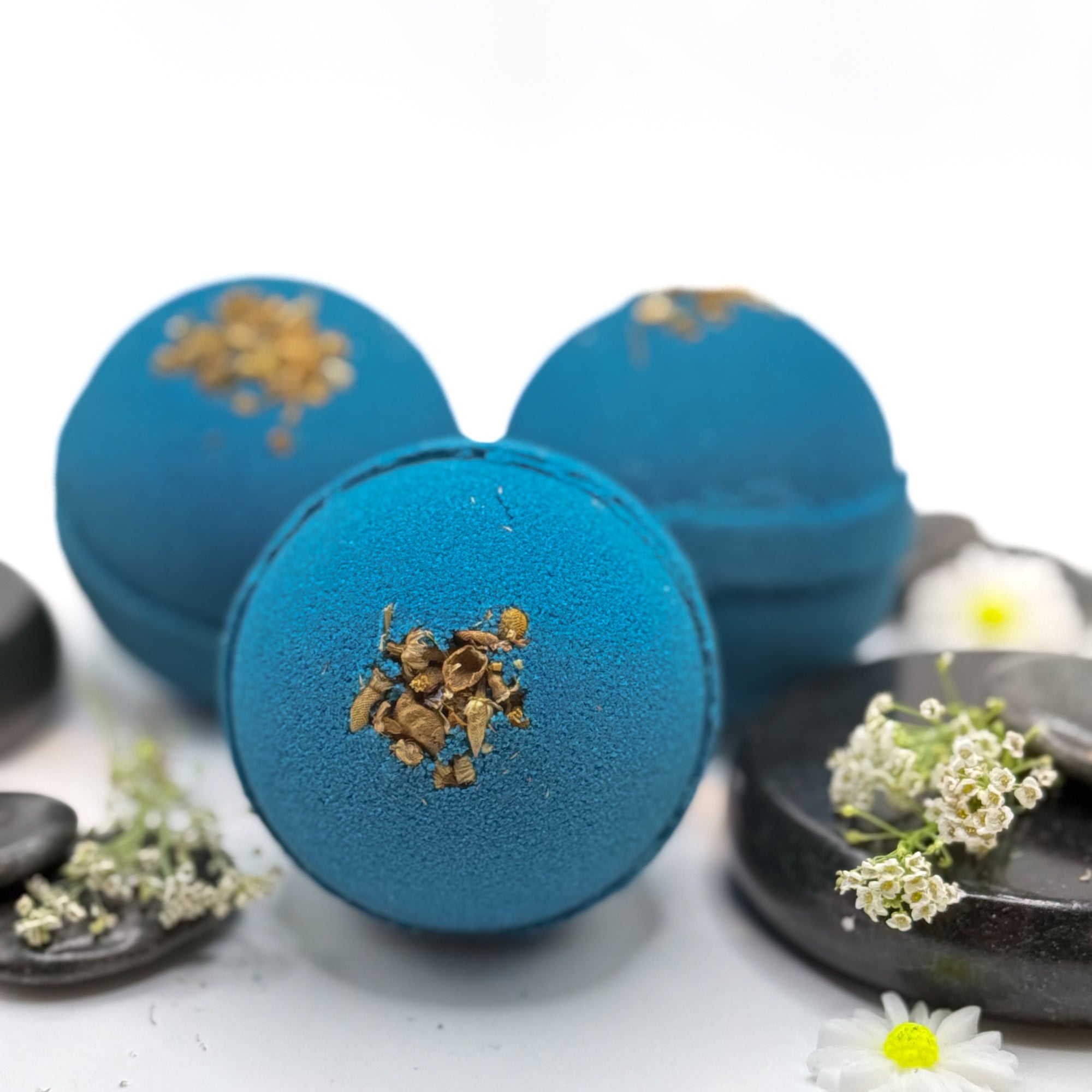 Three blue tranquility bath bombs with dried flower details on a white background with spa type props with a slight blur on the two bath bombs on the background.