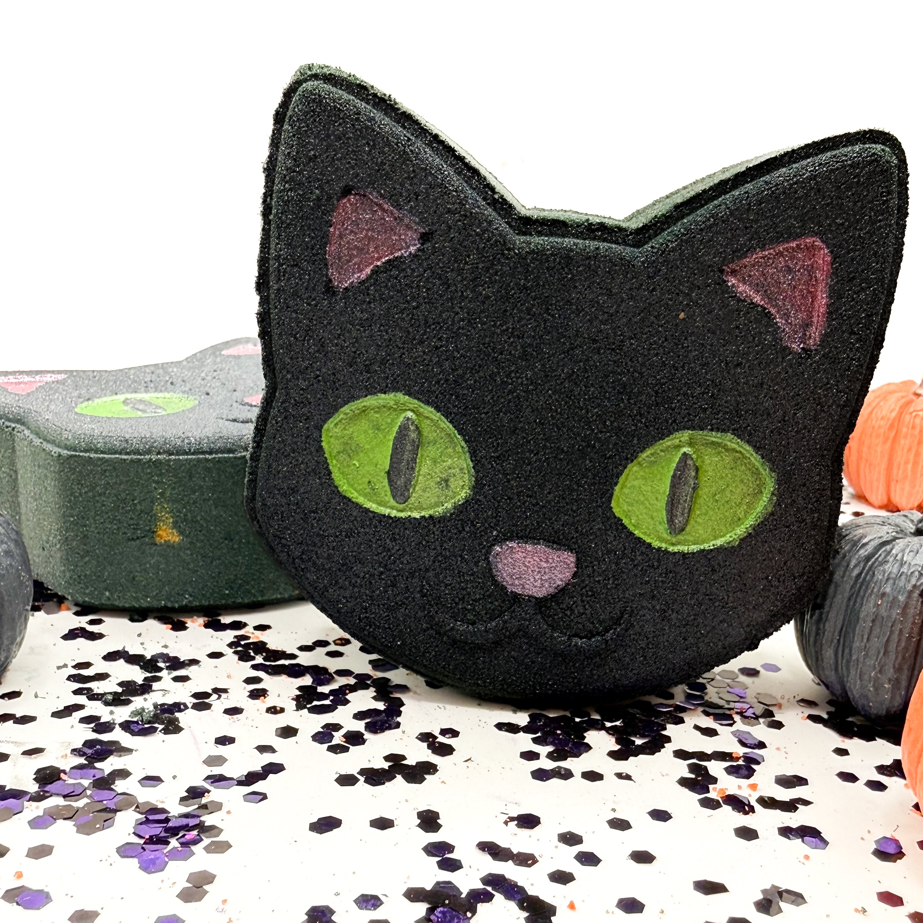 A black cat-shaped bath bomb with green eyes and pink nose, displayed on a white surface scattered with purple and black glitter, with other Halloween themed soaps in the background.