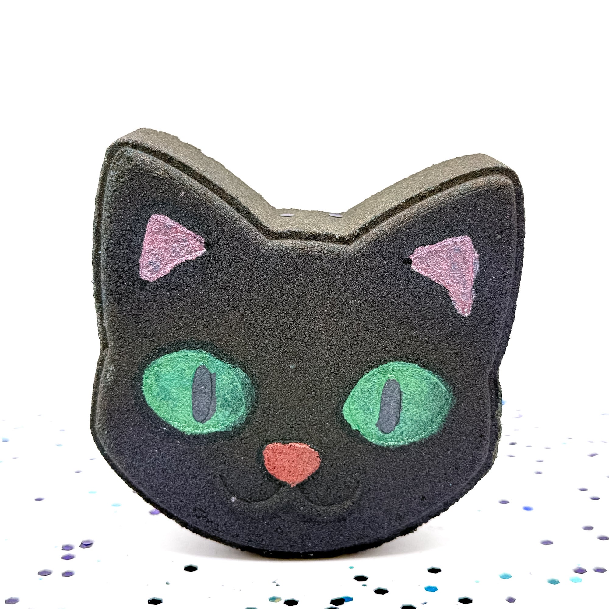 Rusted Acre Soap Co.’s handcrafted Black Cat Bath Bomb standing upright, shaped like a cat’s face with bright green eyes, pink ears, and a red nose, shown against a white background with scattered blue and purple confetti.
