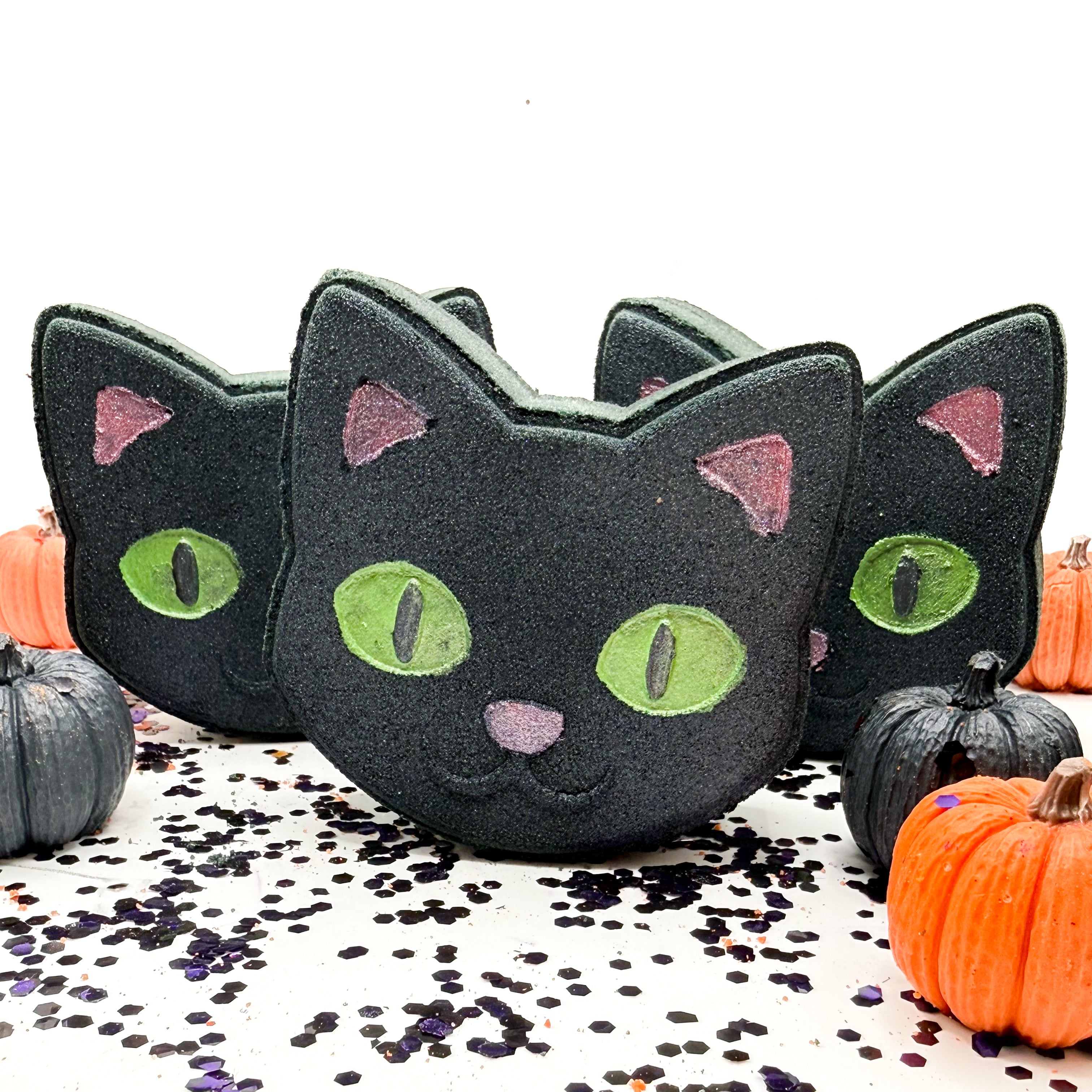 Three black cat shaped bath bombs with hand painted green eyes and pink ears, arranged on a white surface with scattered confetti and small pumpkins.