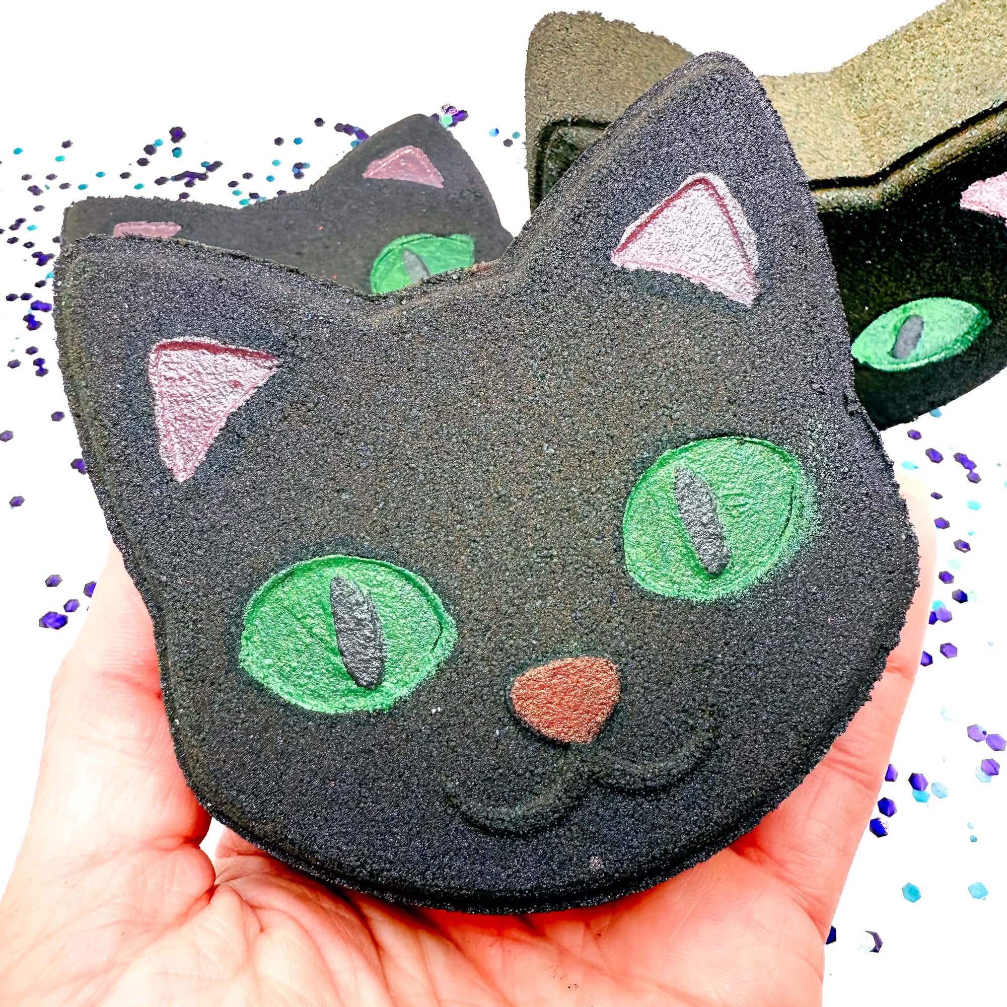 Hand holding Rusted Acre Soap Co.’s Black Cat Bath Bomb with green eyes and pink ears — a spooky, kid-friendly Halloween bath treat.