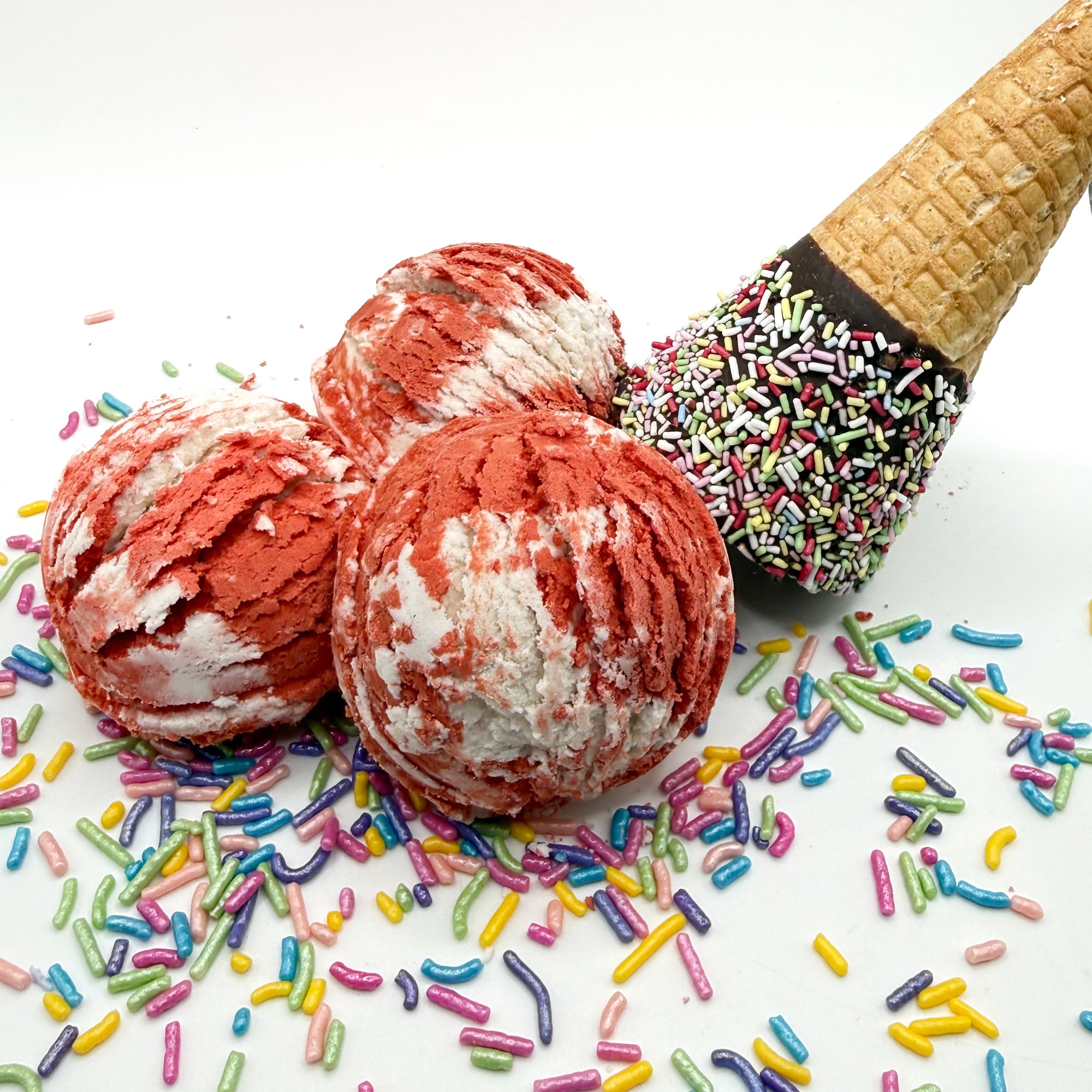 Three red and white cherry scented bubble bath scoops handmade by Rusted Acre Soap Co. Scoop looks like an ice cream scoop. White background with sprinkles and ice cream cone prop on a white background.