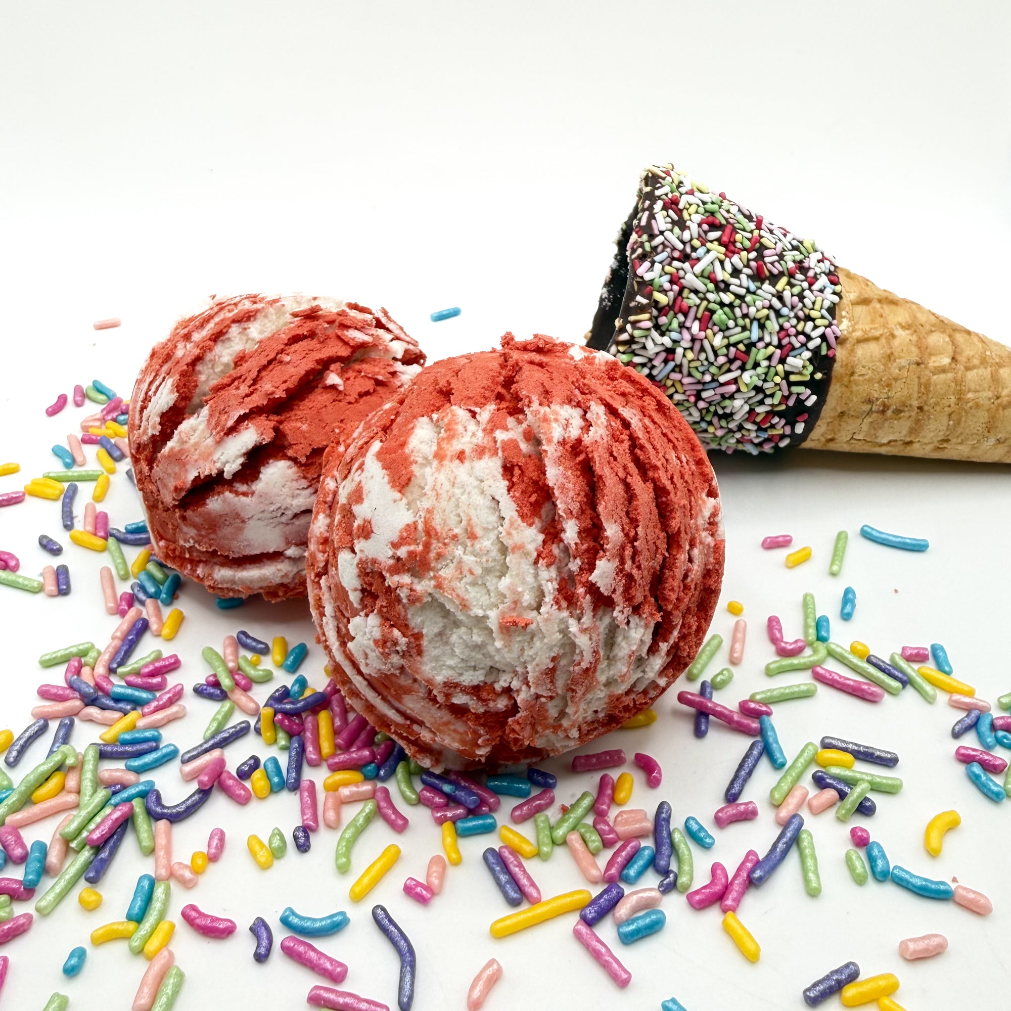 Two red and white cherry scented bubble bath scoop handmade by Rusted Acre Soap Co. Scoop looks like an ice cream scoop. White background with sprinkles and ice cream cone prop on a white background.