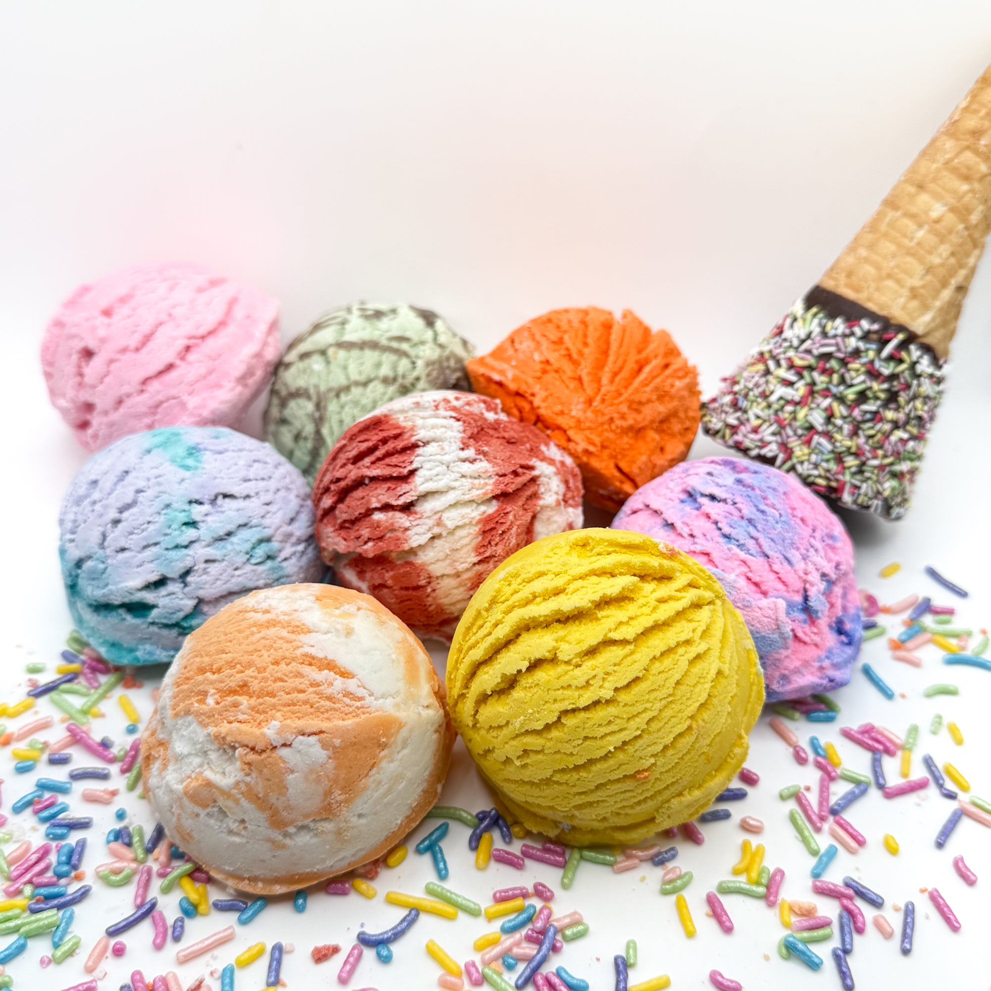 Multiple fun colored and scented bubble bath scoops handmade by Rusted Acre Soap Co. Scoop looks like an ice cream scoop. White background with sprinkles and ice cream cone prop on a white background.