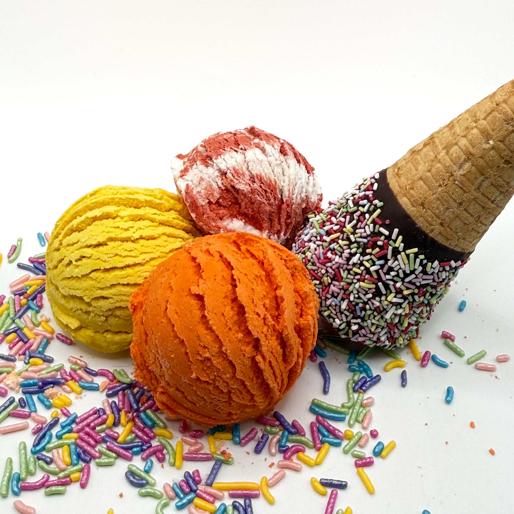 Three different colored and scented bubble bath scoops handmade by Rusted Acre Soap Co. Scoop looks like an ice cream scoop and are orange, yellow and red and white. White background with sprinkles and ice cream cone prop on a white background.