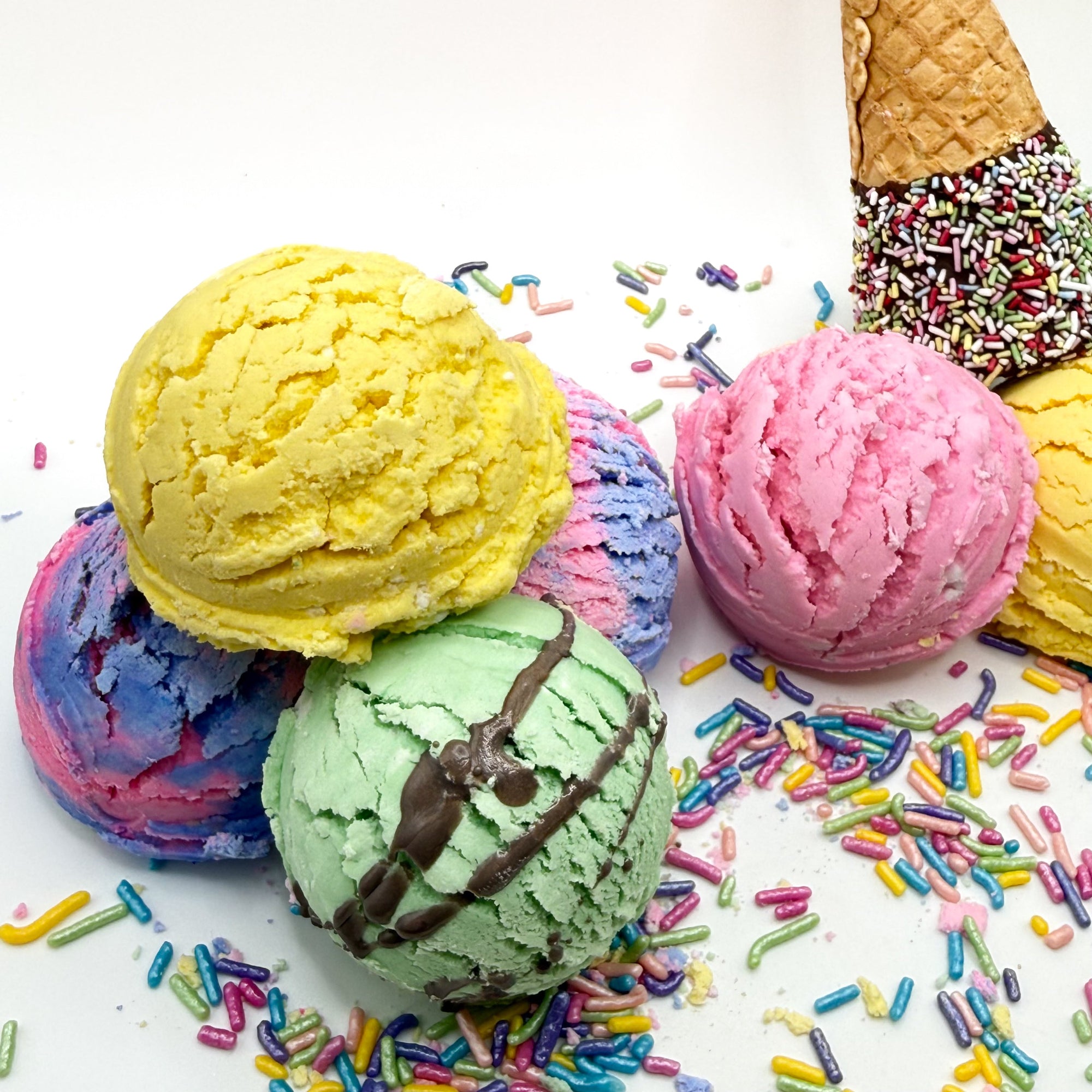 Multiple bubble bath scoops with a chocolate covered cone and rainbow sprinkles handmade by Rusted Acre Soap Co. Colors are yellow, pink, mint green, and 2 are lite purple and pink swirled.