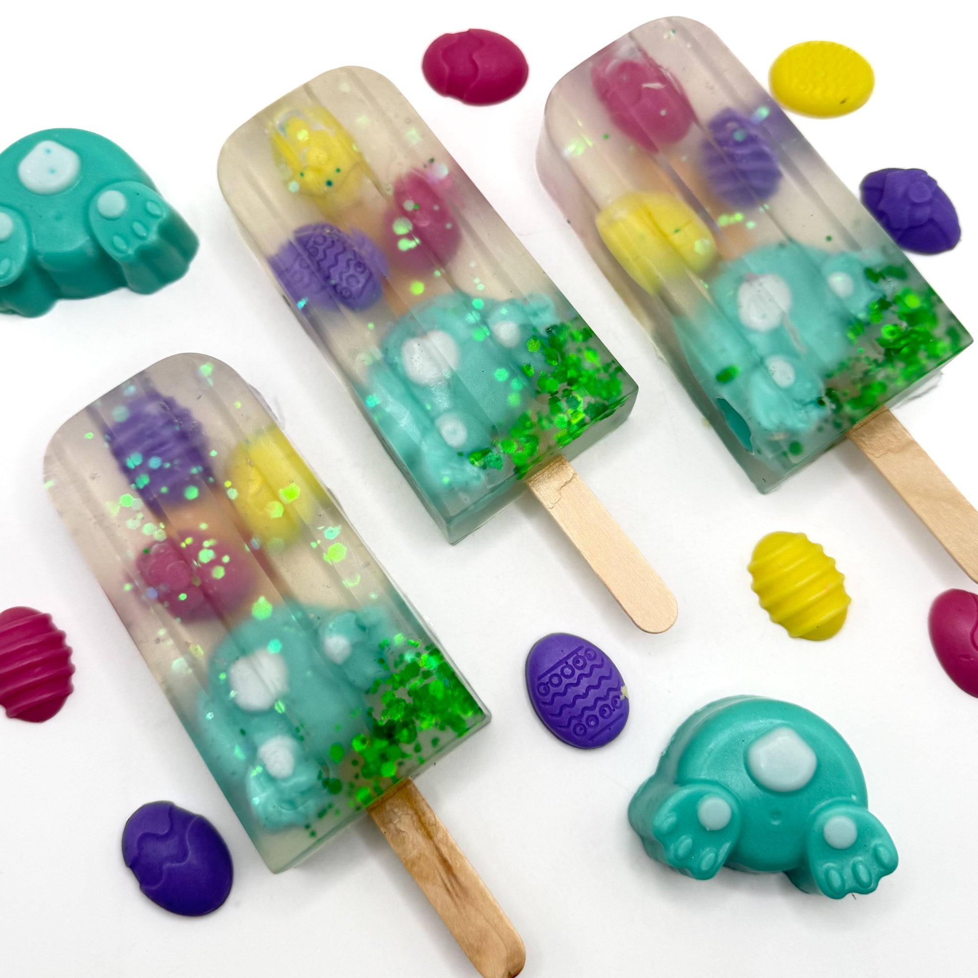 Three Easter popsicle shaped soap bars with a teal bunny butt and egg soaps embedded inside on a white background. Handmade by Rusted Acre Soap Co.