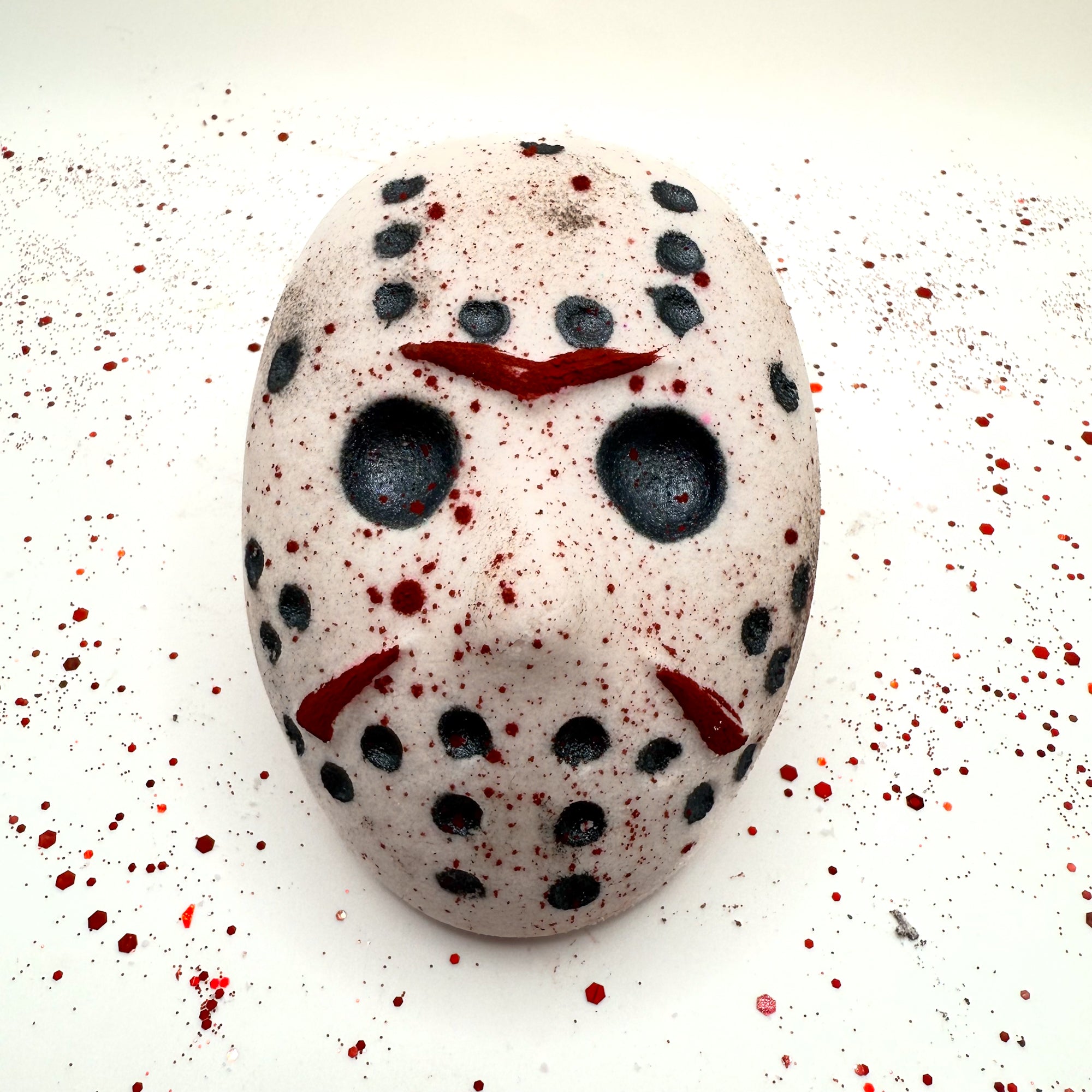 Front view of Camp Slasher horror-inspired bath bomb shaped like a hockey mask, with black and red painted details and blood splatter design.