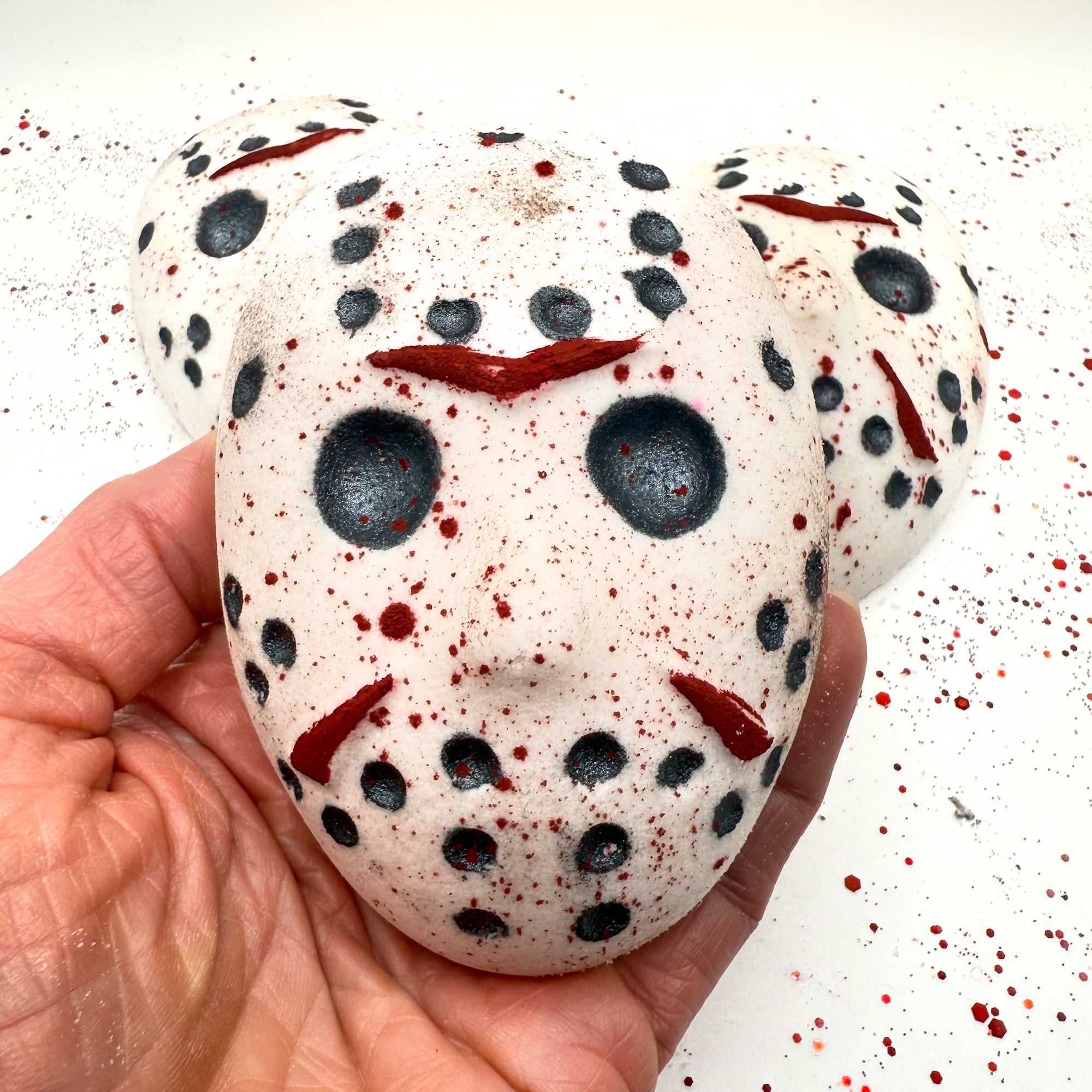 Close-up of Camp Slasher horror bath bomb being held, showing the painted red slashes and black mask holes with splatter design. Made by Rusted Acre Soap Company