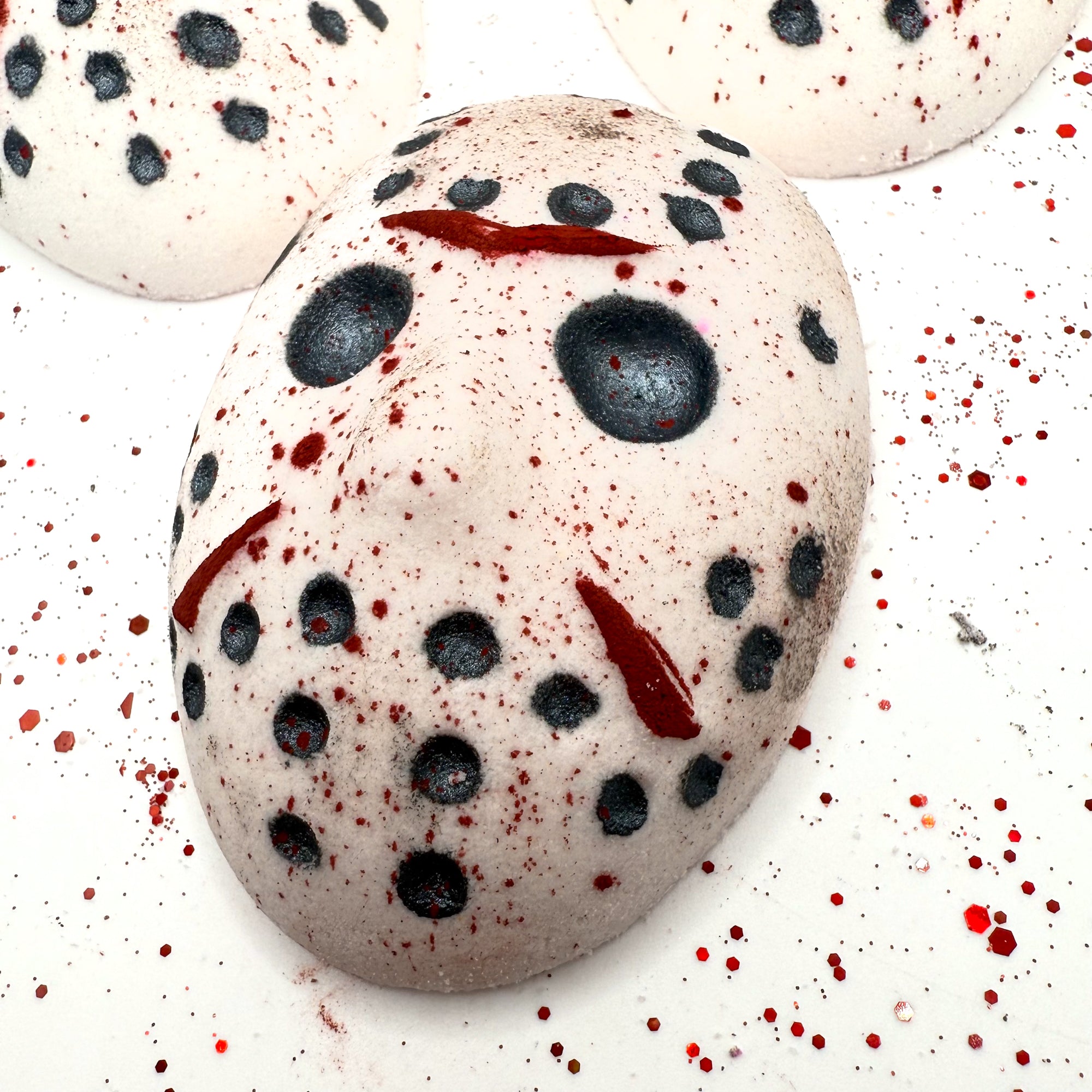 Angled view of Camp Slasher bath bomb laying flat, showcasing detailed red and black painted mask design with scattered blood splatter.