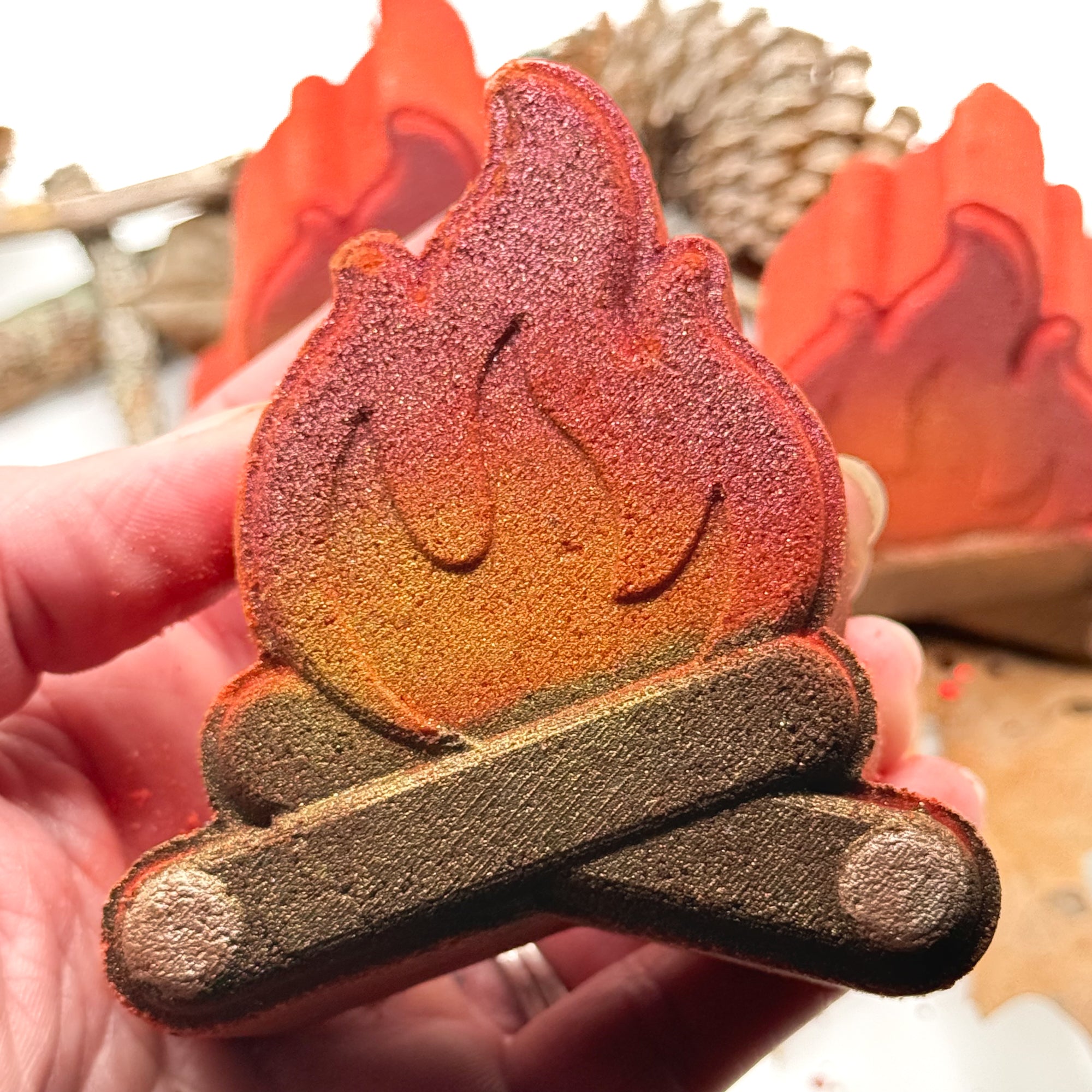 One hand painted campfire bath bomb being held in a hand with leaves, sticks and pinecones on a white background.
