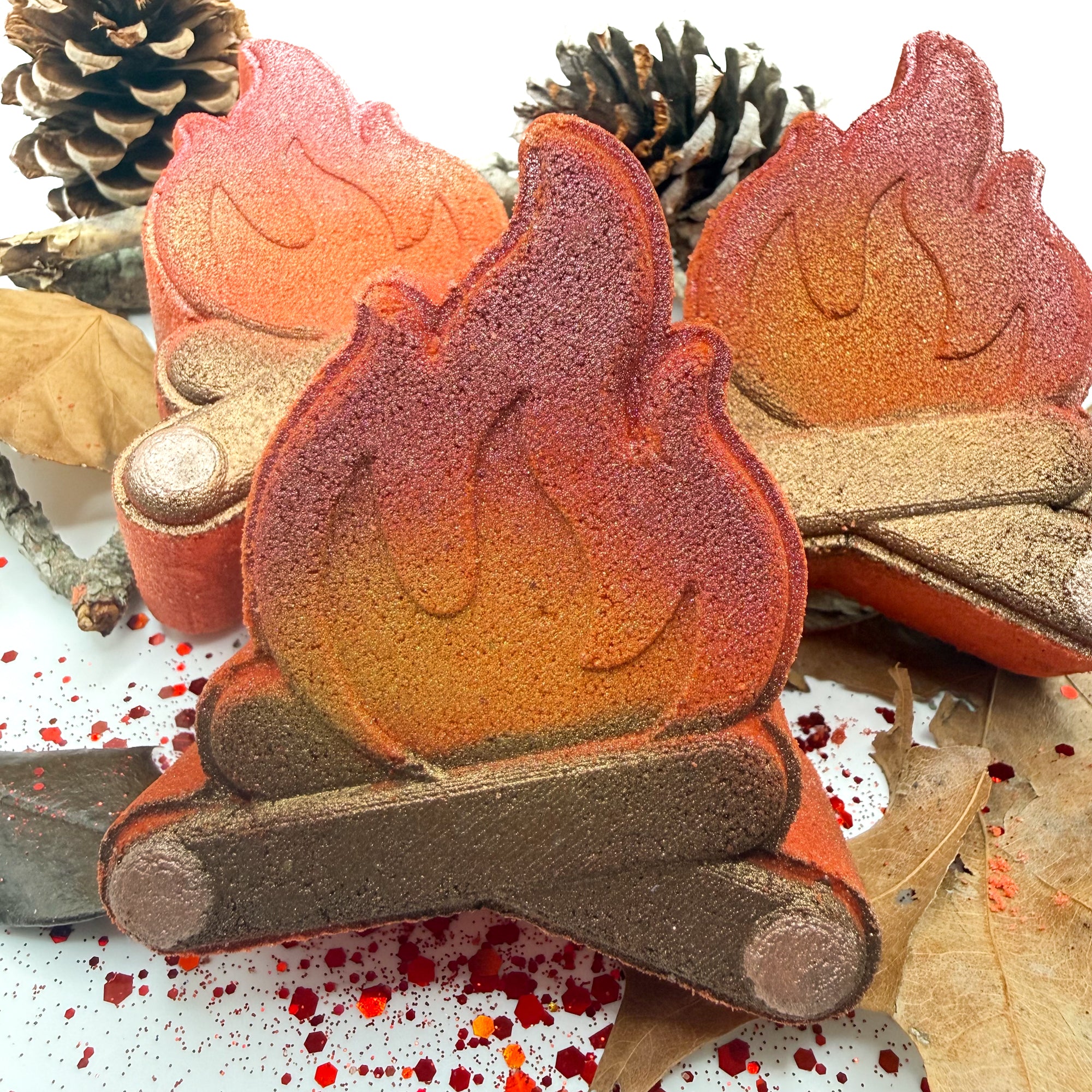 One hand painted campfire bath bomb leaned up against two more campfire bath bombs with leaves, sticks and pinecones on a white background.