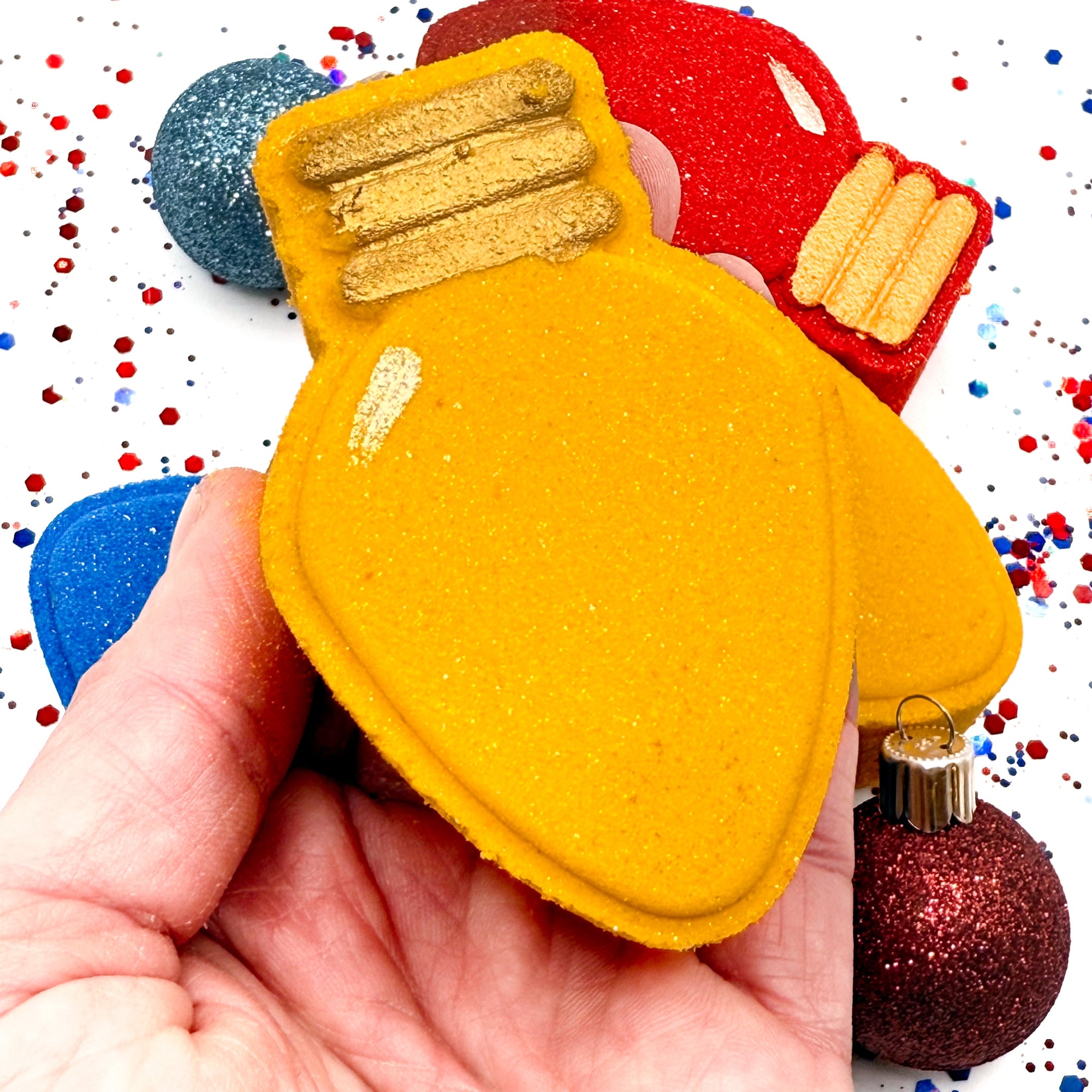 Yellow holiday light bath bomb held in a hand with red, blue, and gold Christmas decorations in the background.