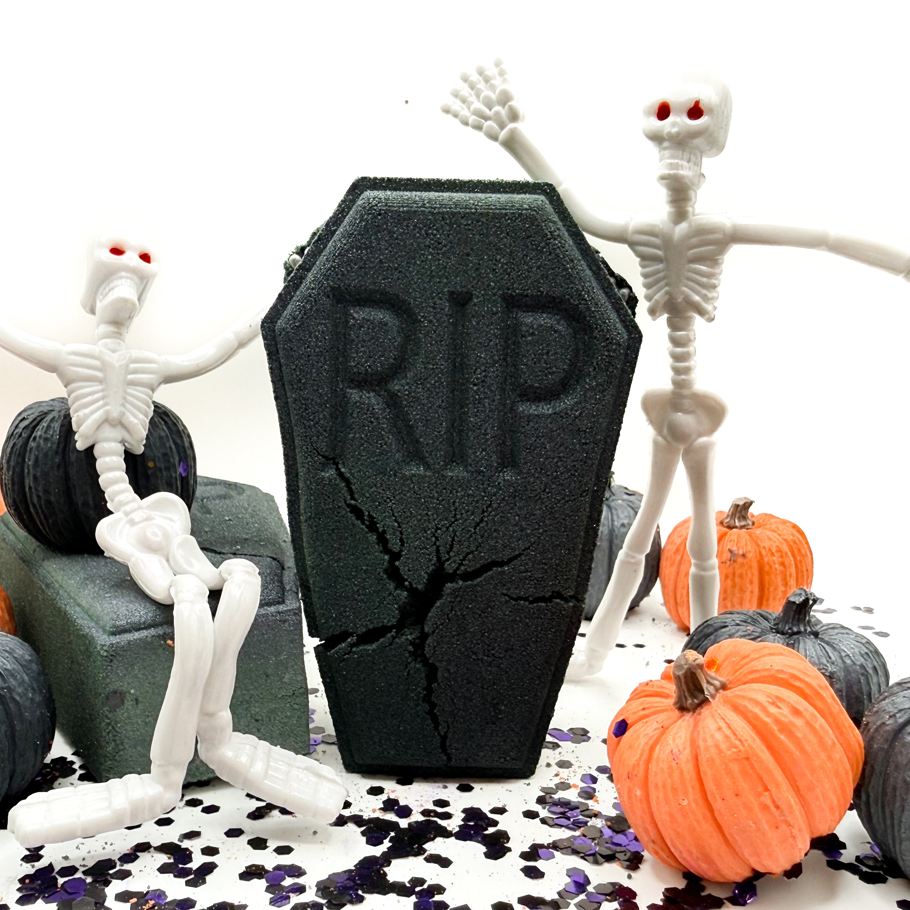 A Halloween themed product display with a black coffin-shaped bath bomb with embossed 'RIP' text and cracks, surrounded by white plastic skeletons, small pumpkins, and confetti on a white background.
