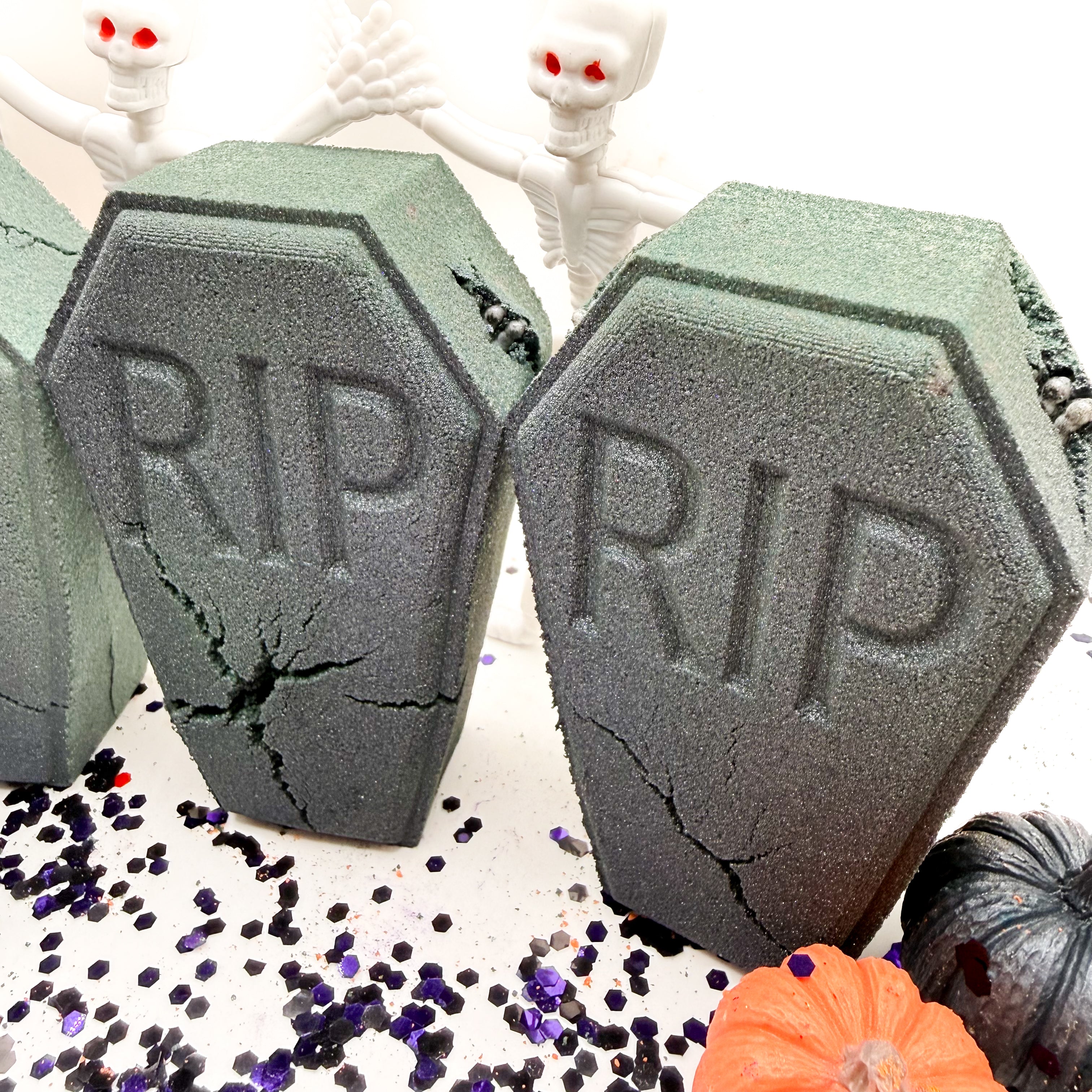 Two gray coffin shaped foam bath bombs with 'RIP' embossed on them, displayed on a white surface with Halloween props including small skeletons, purple glitter, and miniature pumpkins.