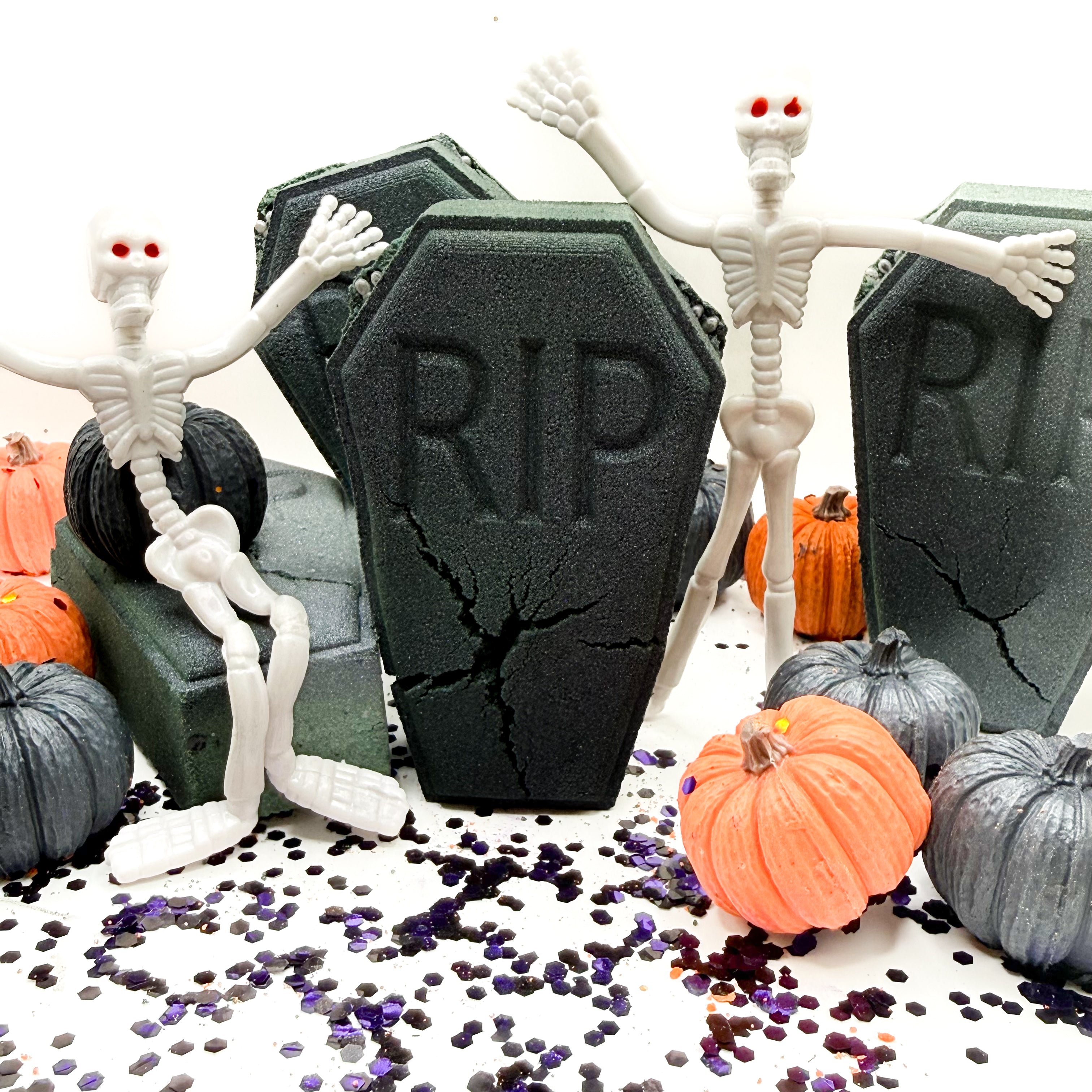 Halloween themed tombstone bath bomb with three white plastic skeleton figures posed around dark gray and black RIP gravestones, alongside small pumpkins and scattered purple and black confetti on a white surface.
