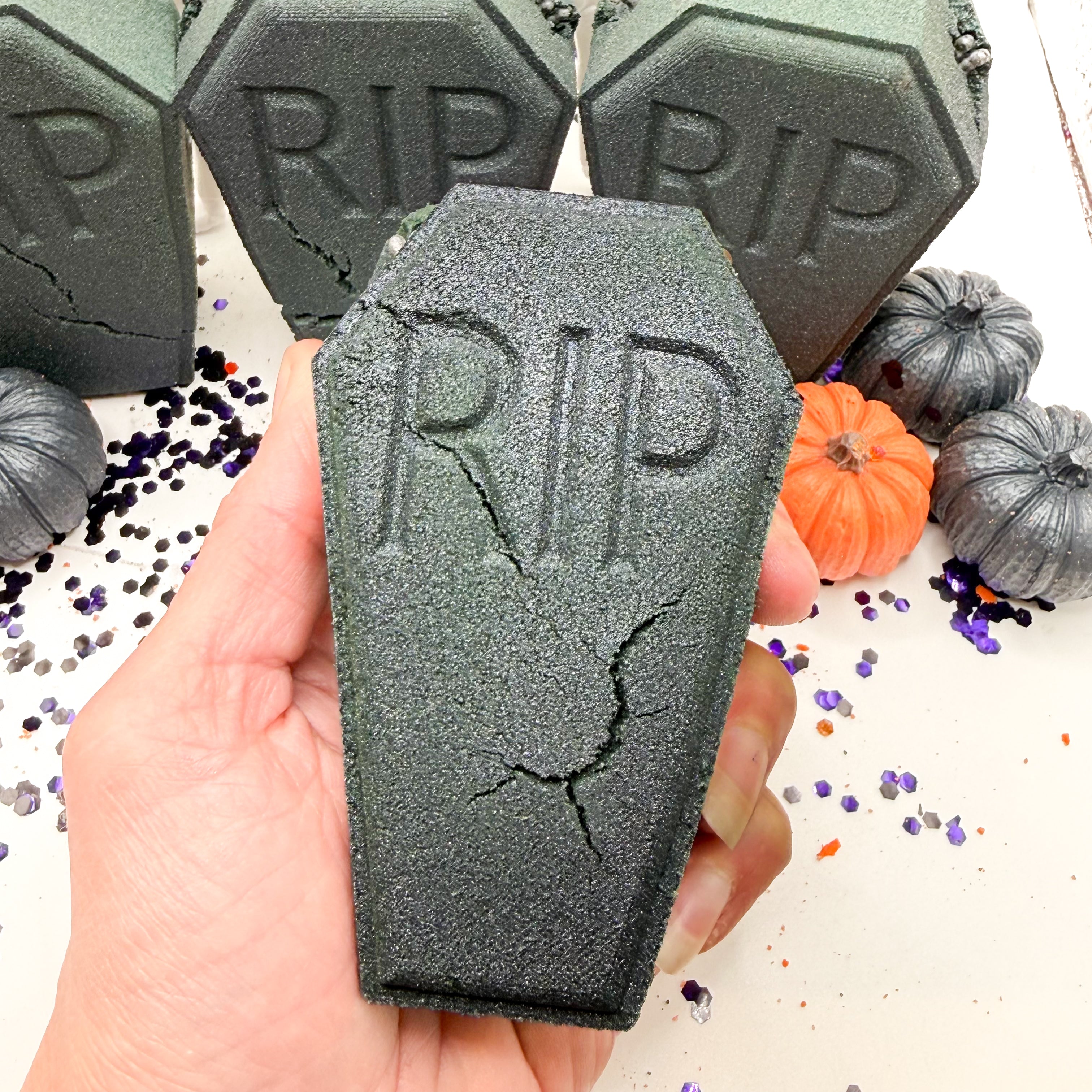 A hand holds a dark gray coffin shaped bath bomb with 'RIP' embossed on it, with more matching coffin-shaped items and small pumpkins in the background.