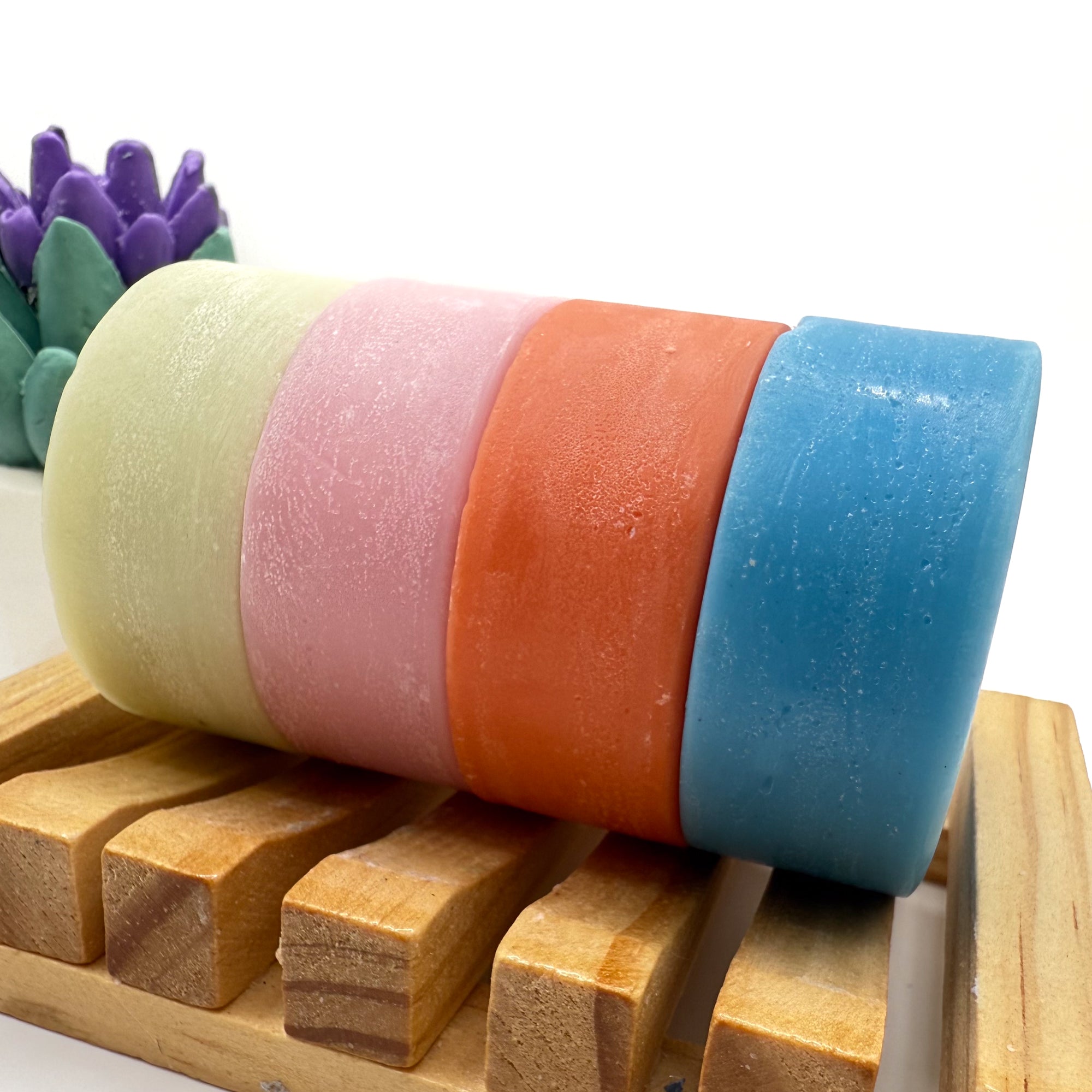 Four solid conditioner bars on a wood soap dish, green, pink, orange and blue.