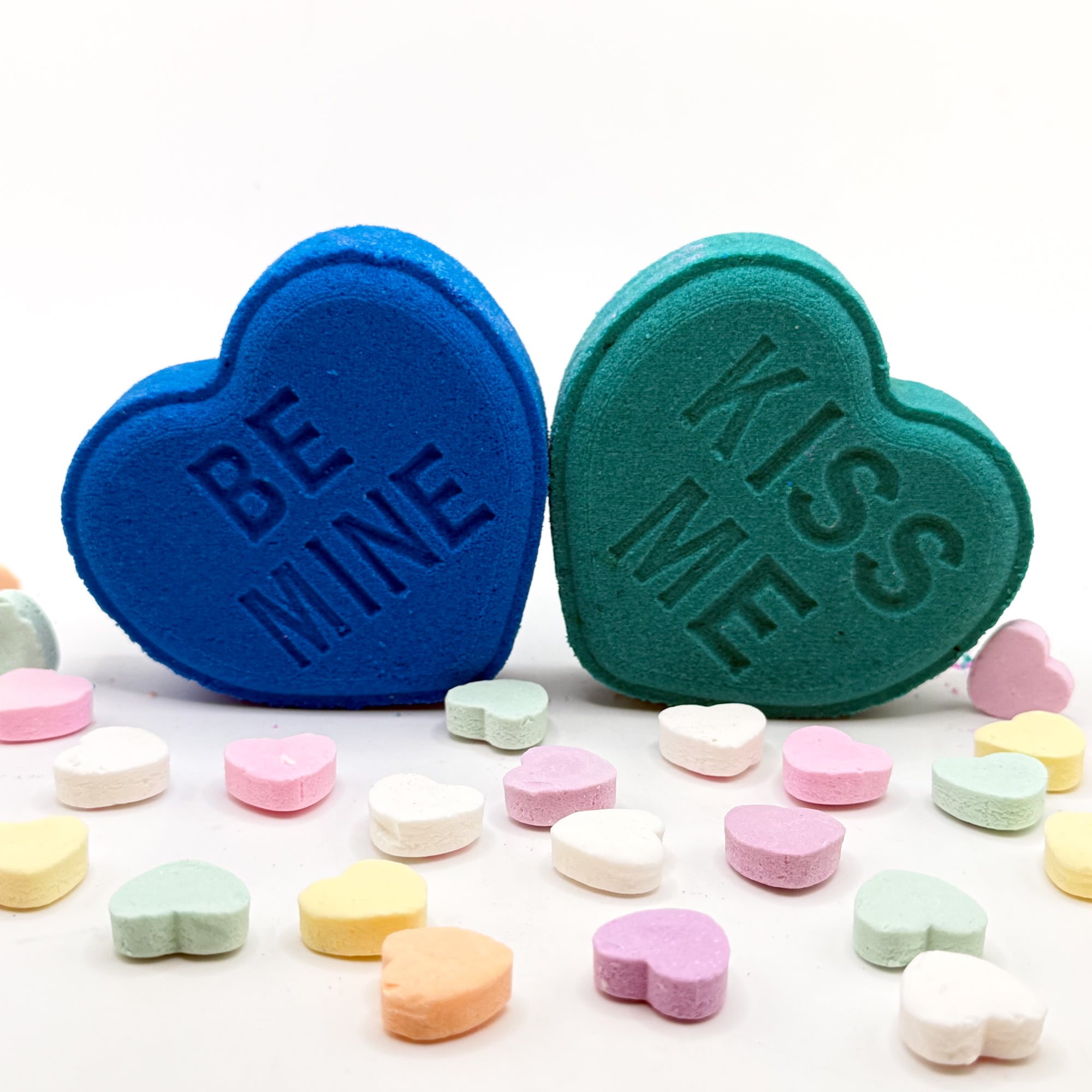 Blue and green heart shaped bath bombs with 'Be Mine' and 'Kiss Me' text on a white background.