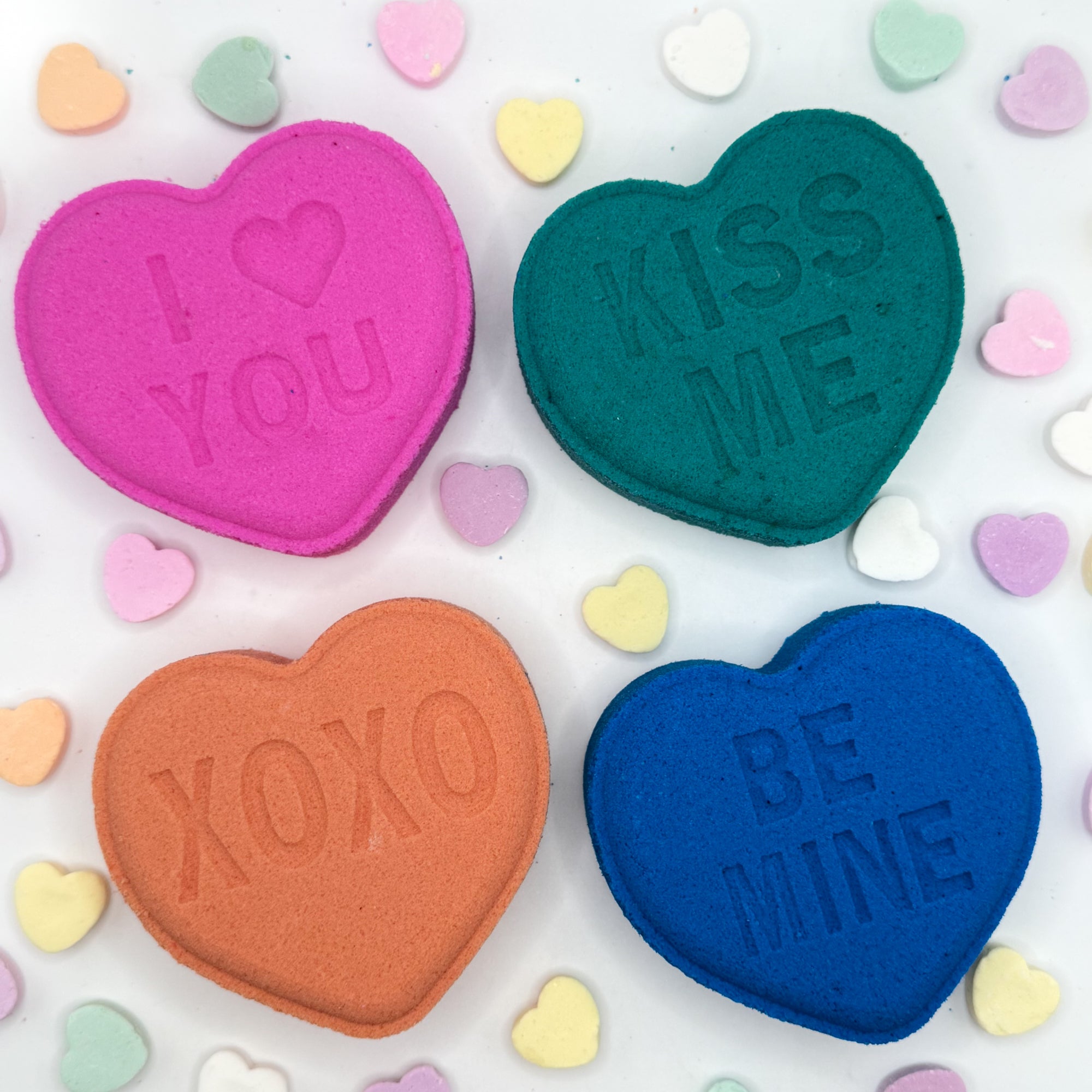 Four heart shaped bath bombs with messages 'I ❤️ YOU', 'KISS ME', 'XOXO', and 'BE MINE' on a colorful heart-patterned background.