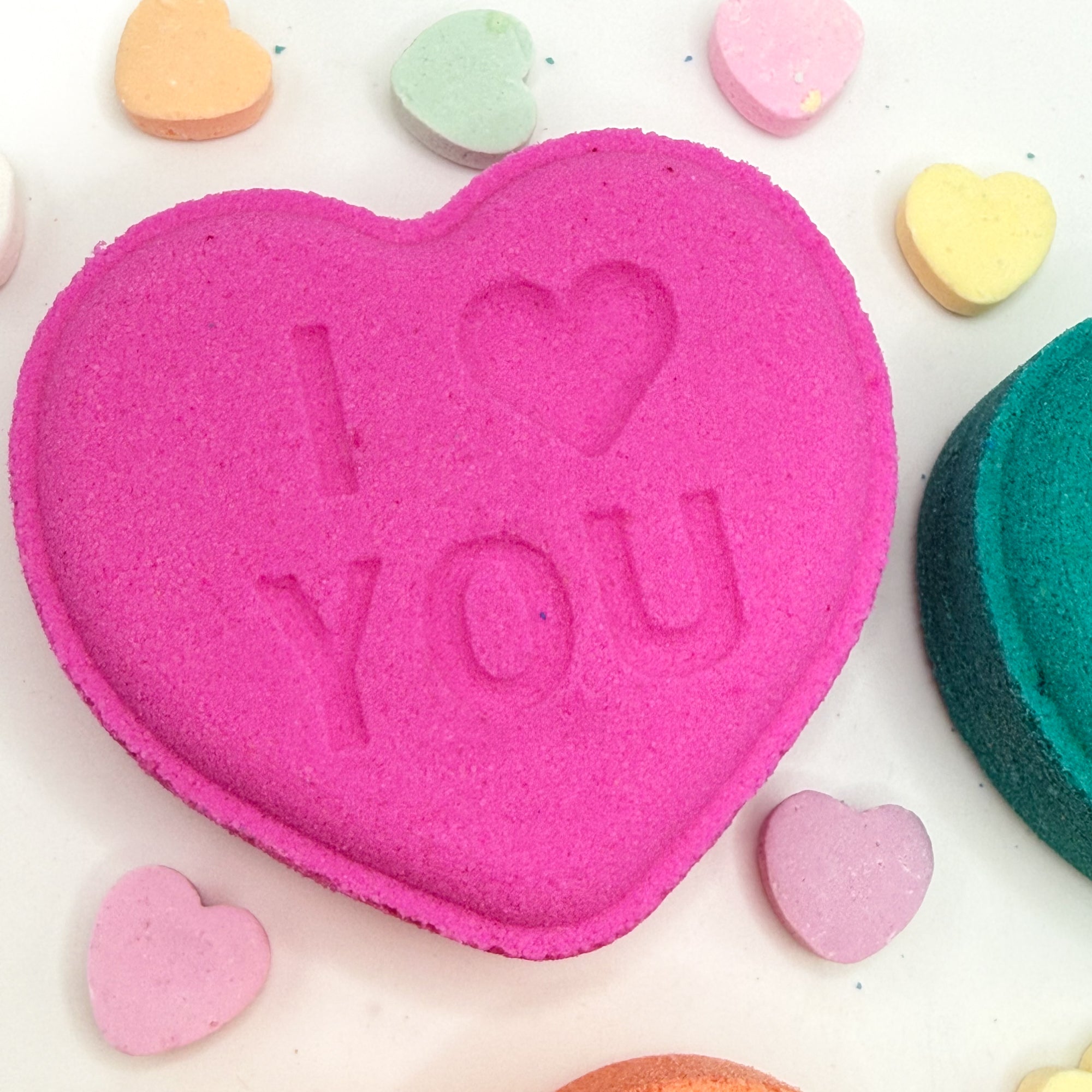 Pink heart shaped bath bomb with 'I love you' text surrounded by colorful heart-shaped candies on a white background