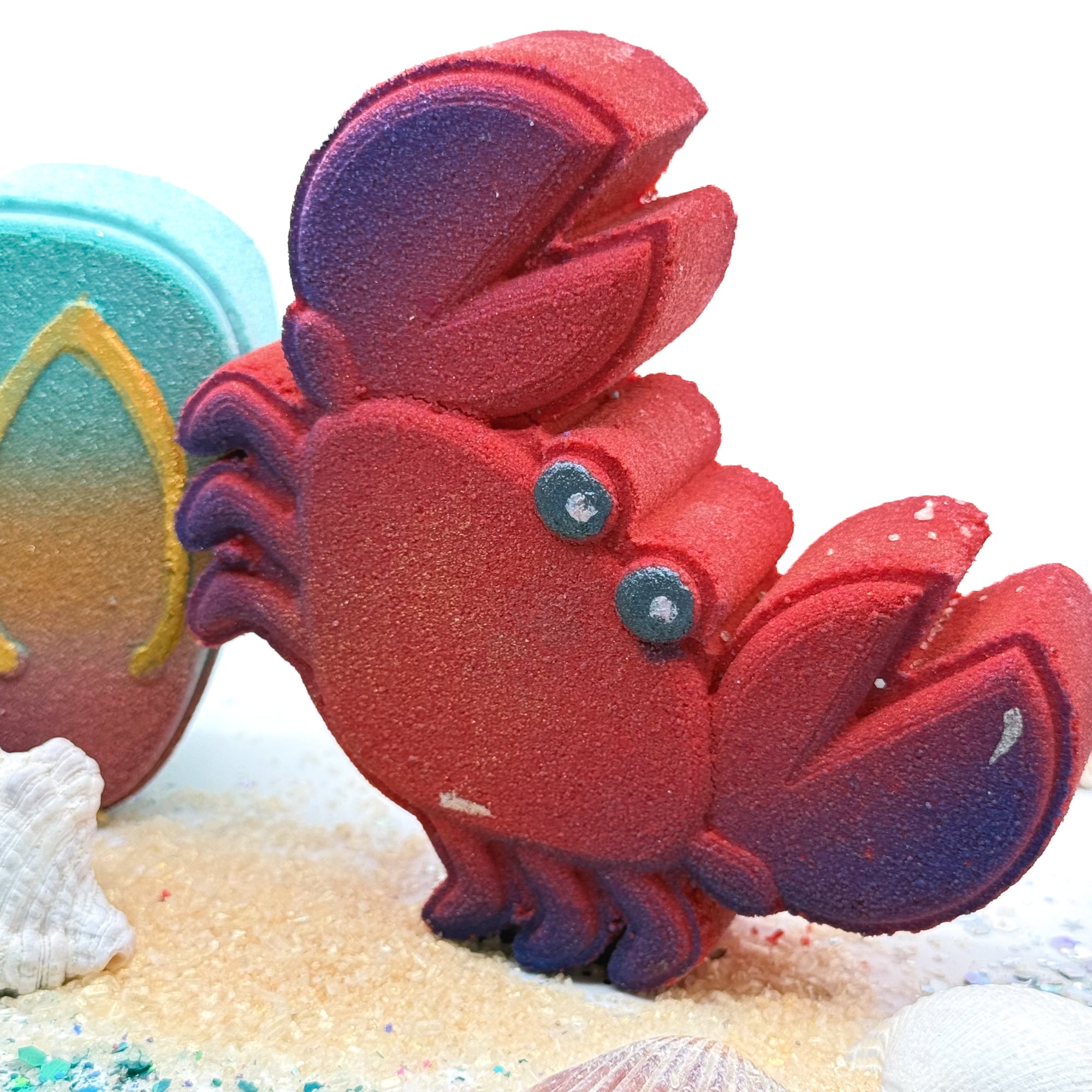 Red crab bath bomb on a sandy surface with shells in the background