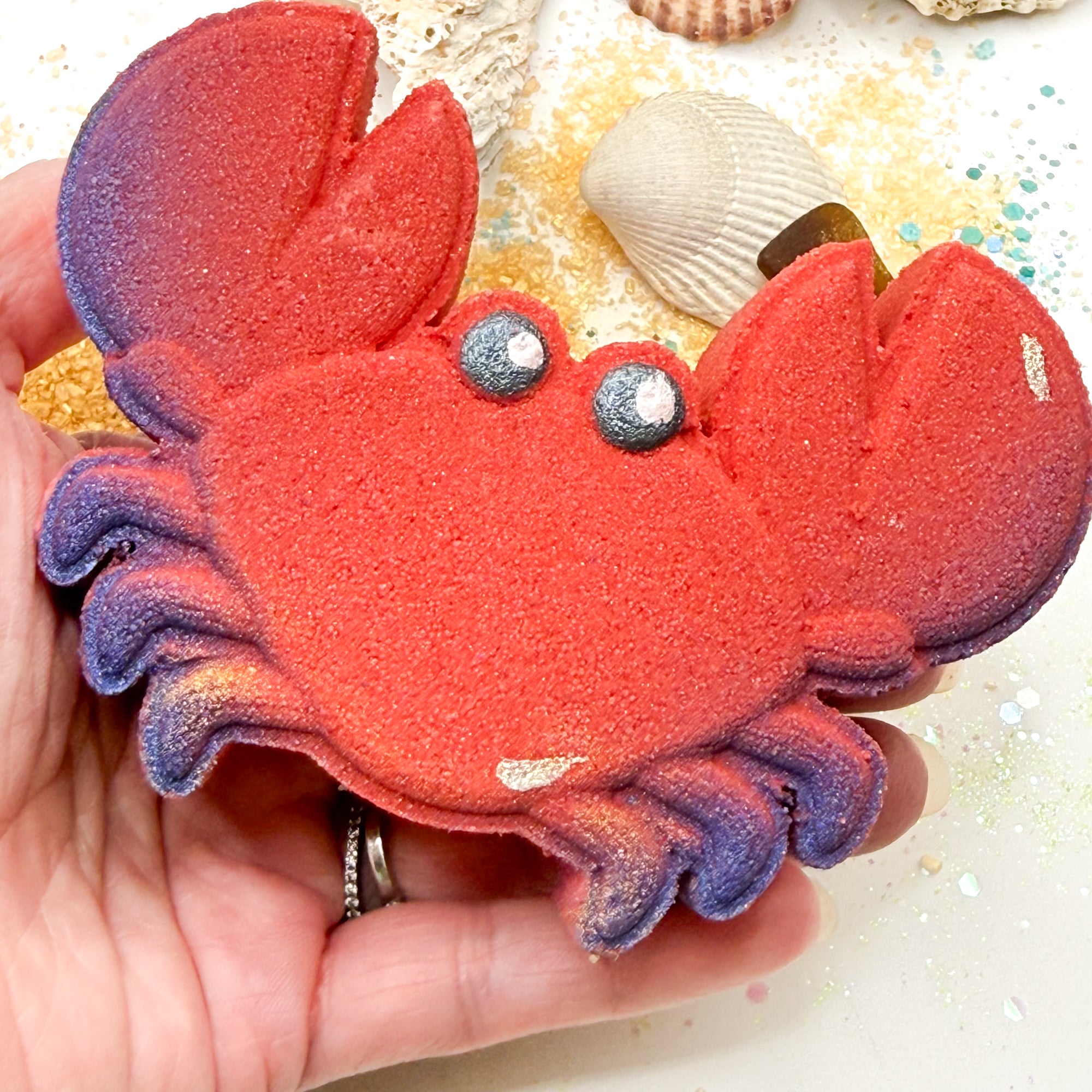 Red crab bath bomb with blue pinchers being held in a hand over a beach themed background.