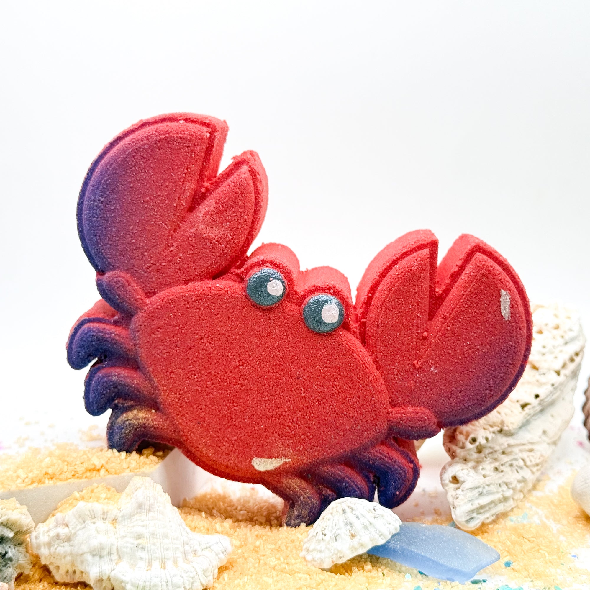 Handmade red crab bath bomb in sand displayed with shells and sand.