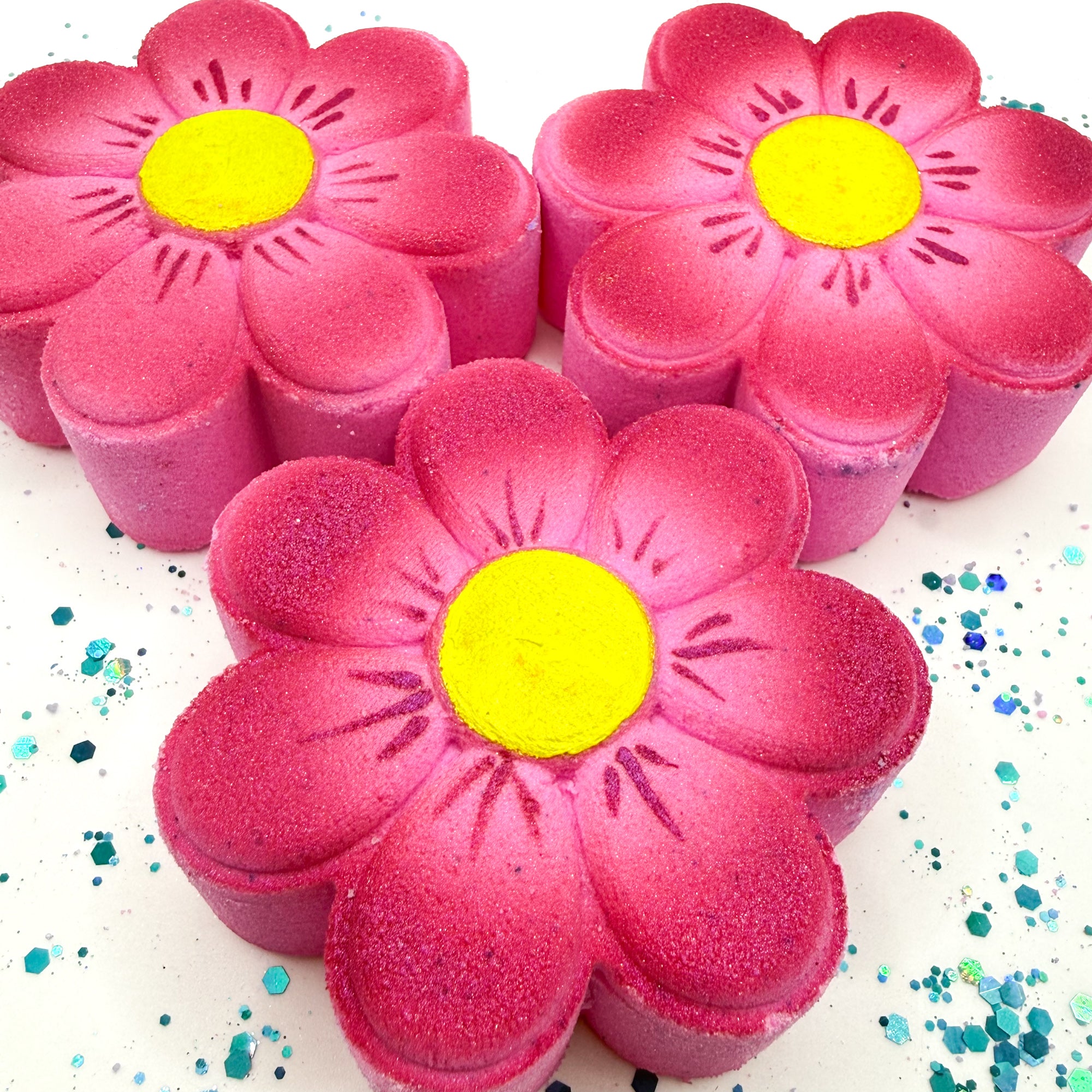 Pink daisy bath bombs with yellow centers on a white background with blue and green confetti.
