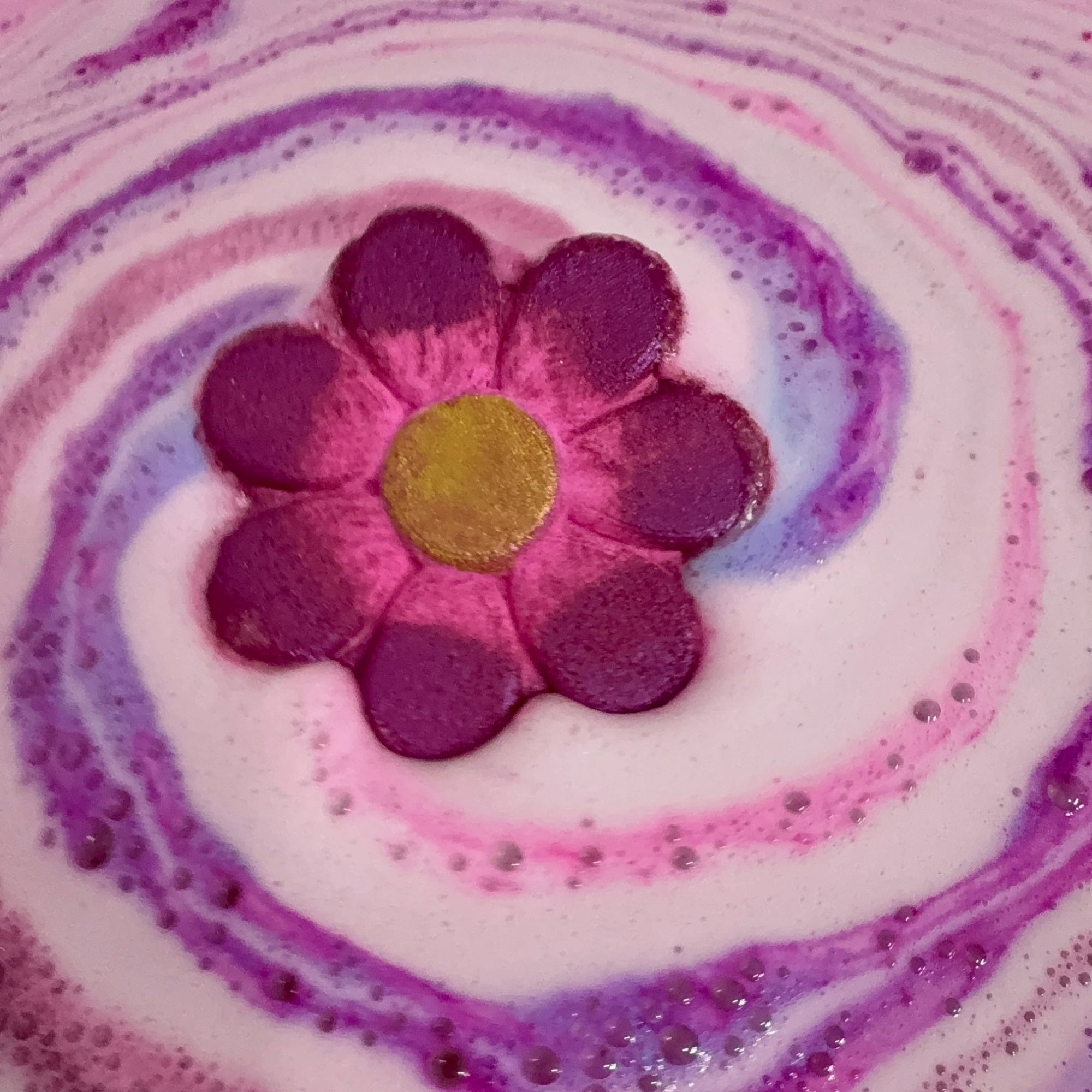 Rusted Acre Soap Companies daisy bath bomb in the water with pink and purple swirls.