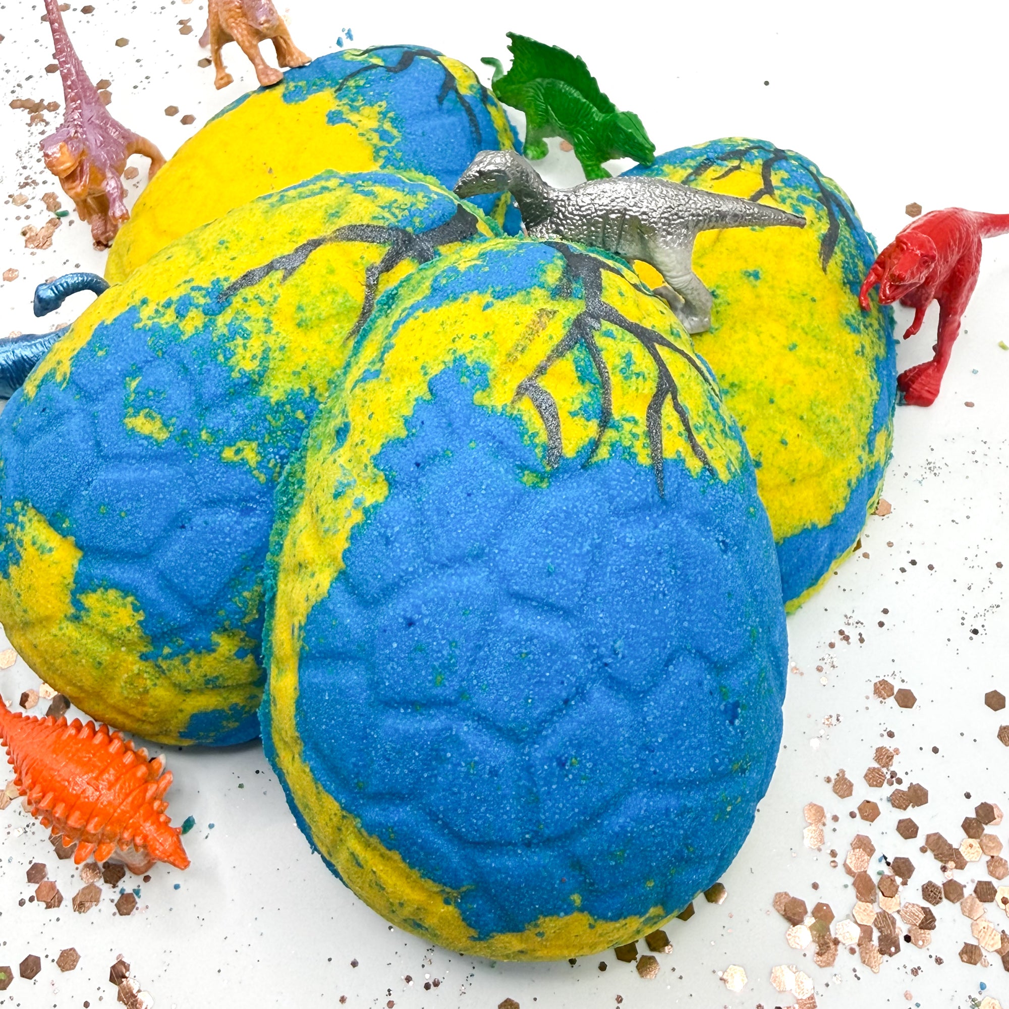 Blue and yellow dinosaur egg bath bombs with toy dinosaurs on a white background