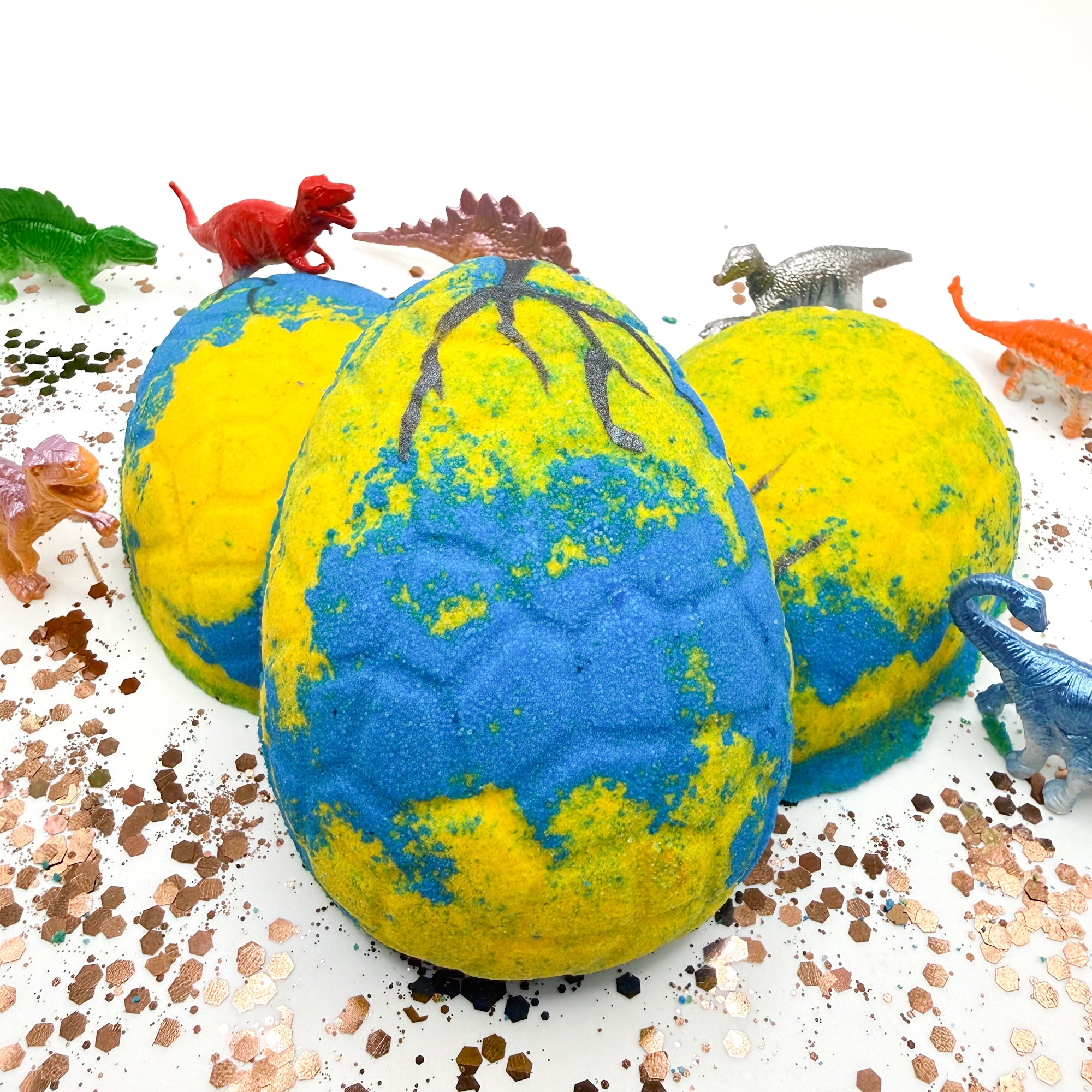 Dinosaur egg bath bombs with blue and yellow pattern, surrounded by small dinosaur figurines that are inside the bath bomb on a white background.