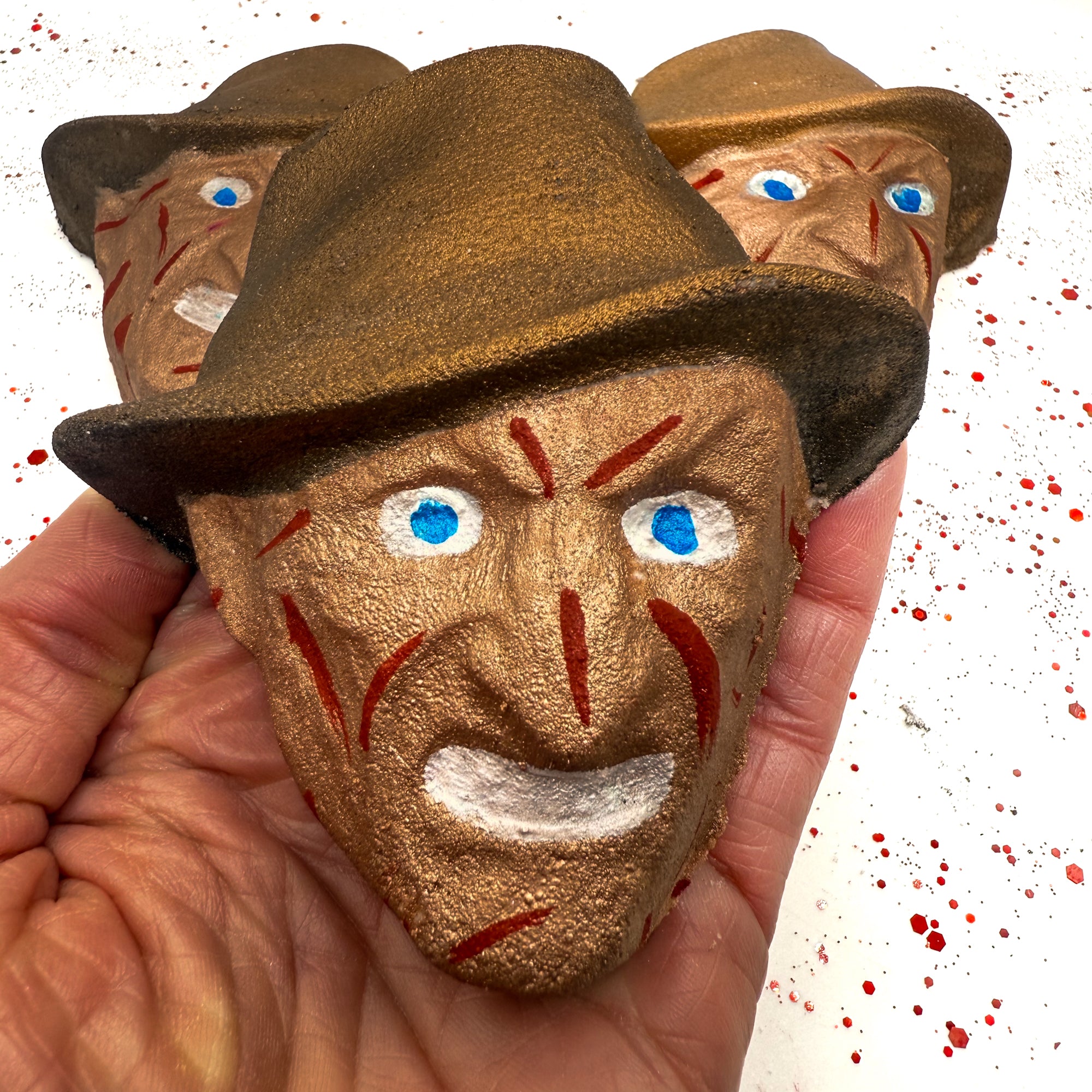 Close-up of Dream Hunter horror movie-inspired bath bomb held in hand, showing detailed hand-painted facial scars, hat, and bright blue eyes.