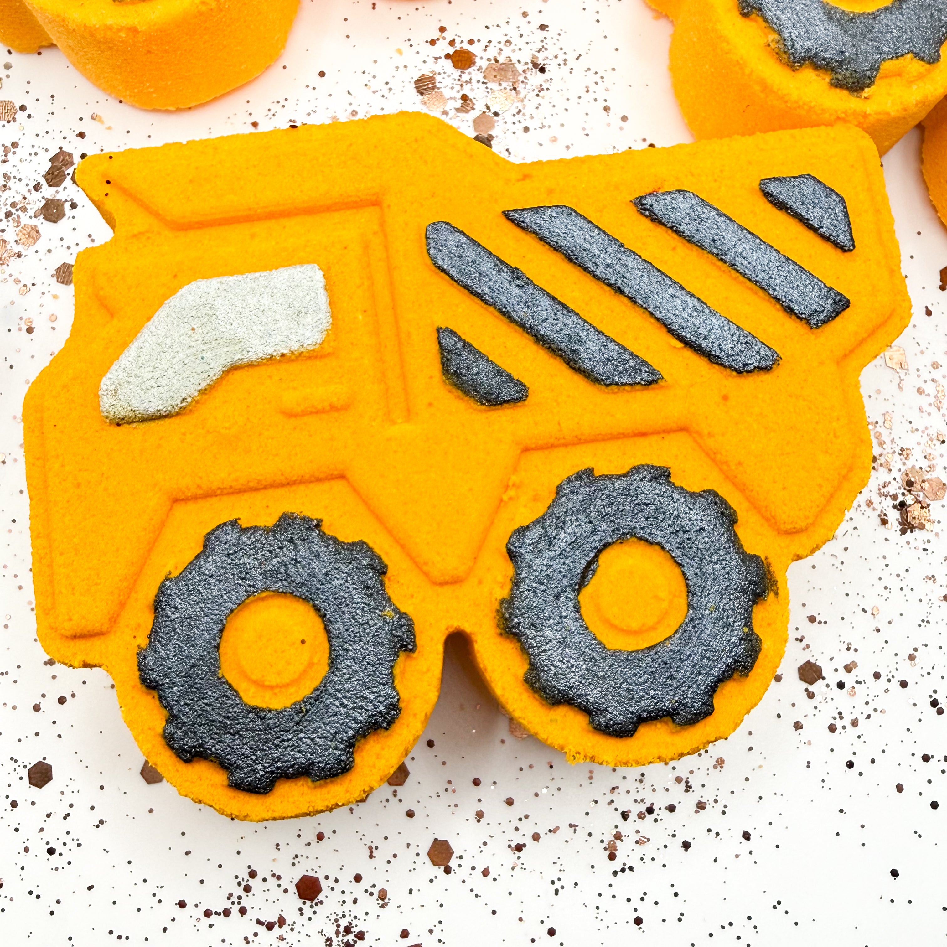 A bright yellow dump truck bath bomb shown on a white speckled surface with other similar orange shapes partially visible in the background.