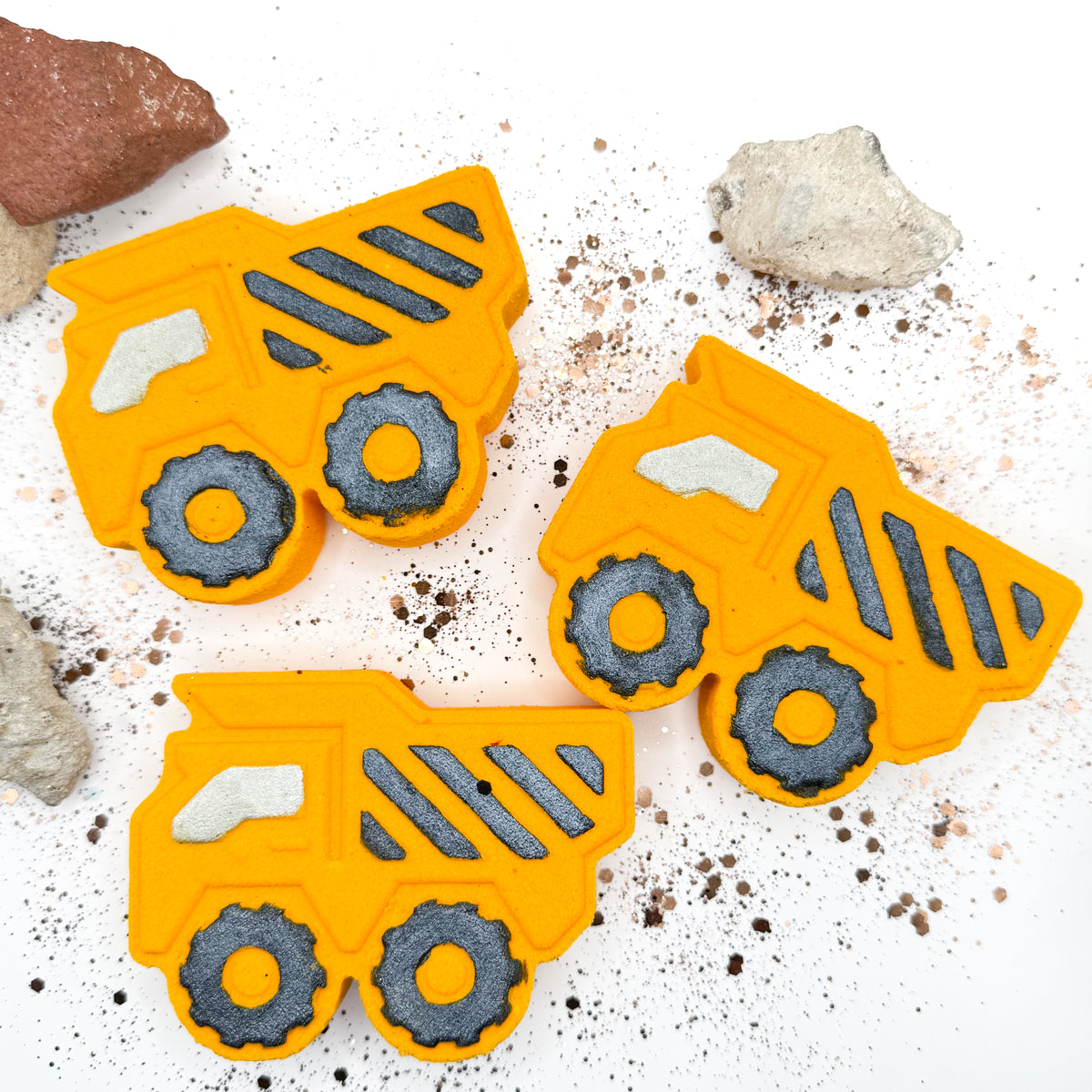 Three bright orange dump truck bath bombs arranged on a white surface with scattered dark specks and small rocks. The items have dark gray wheel details and light gray window accents.