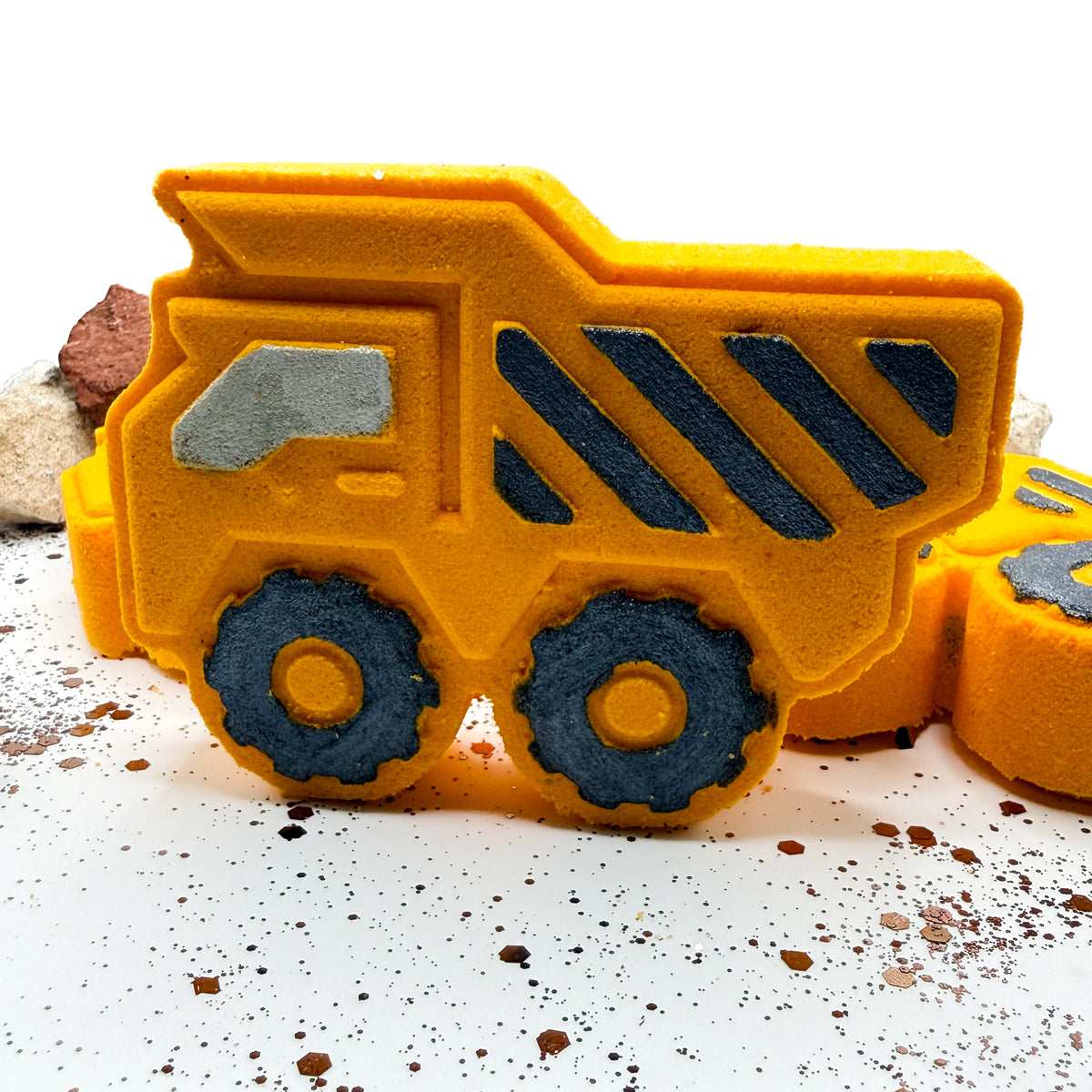 A yellow dump truck shaped bath bomb with hand painted black wheels and gray window details, shown on a white surface with small construction-themed props.
