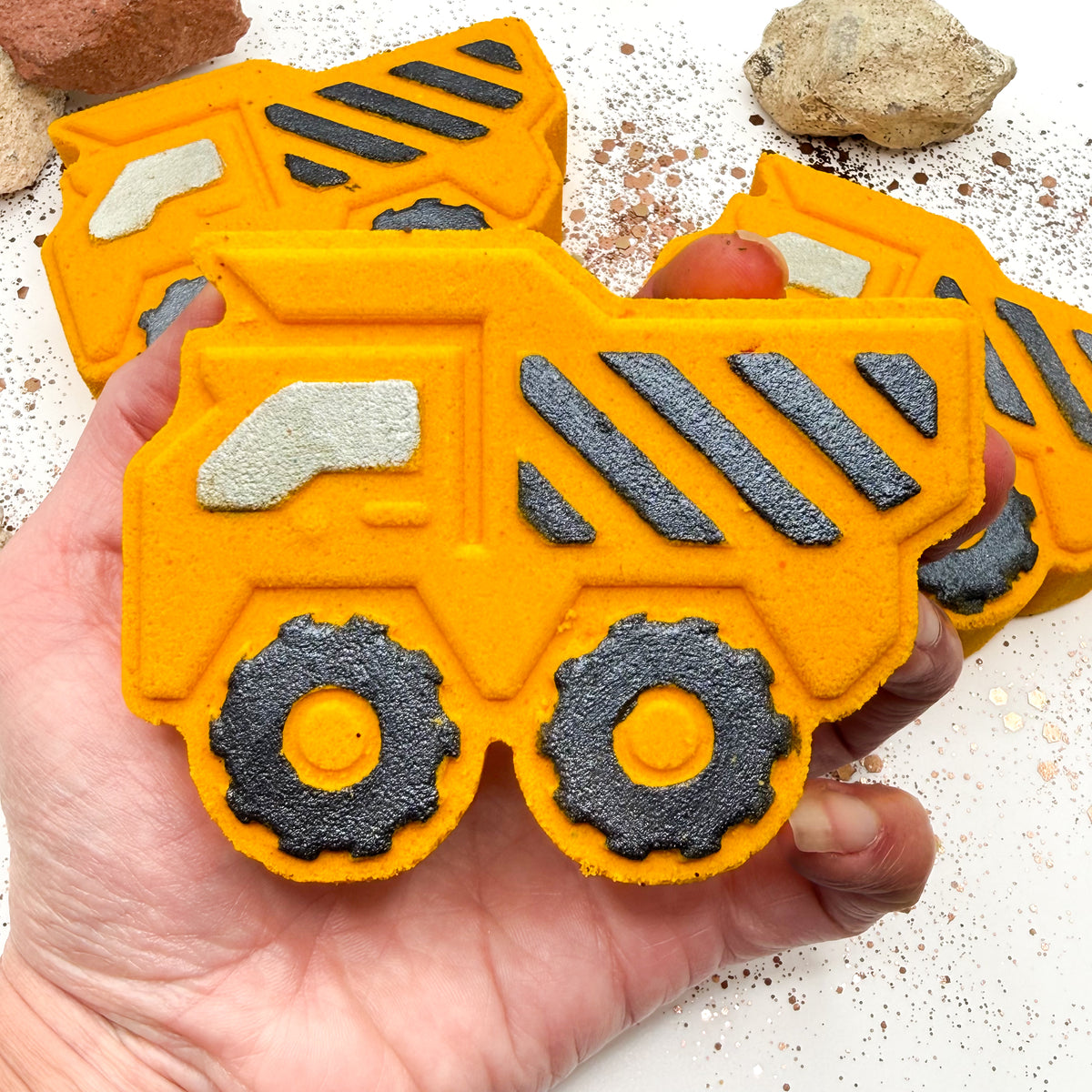 A hand holds a yellow dump truck shaped bath bomb, with black wheels and striped detailing. More matching yellow dump truck items are visible in the background on a light surface with small stones.