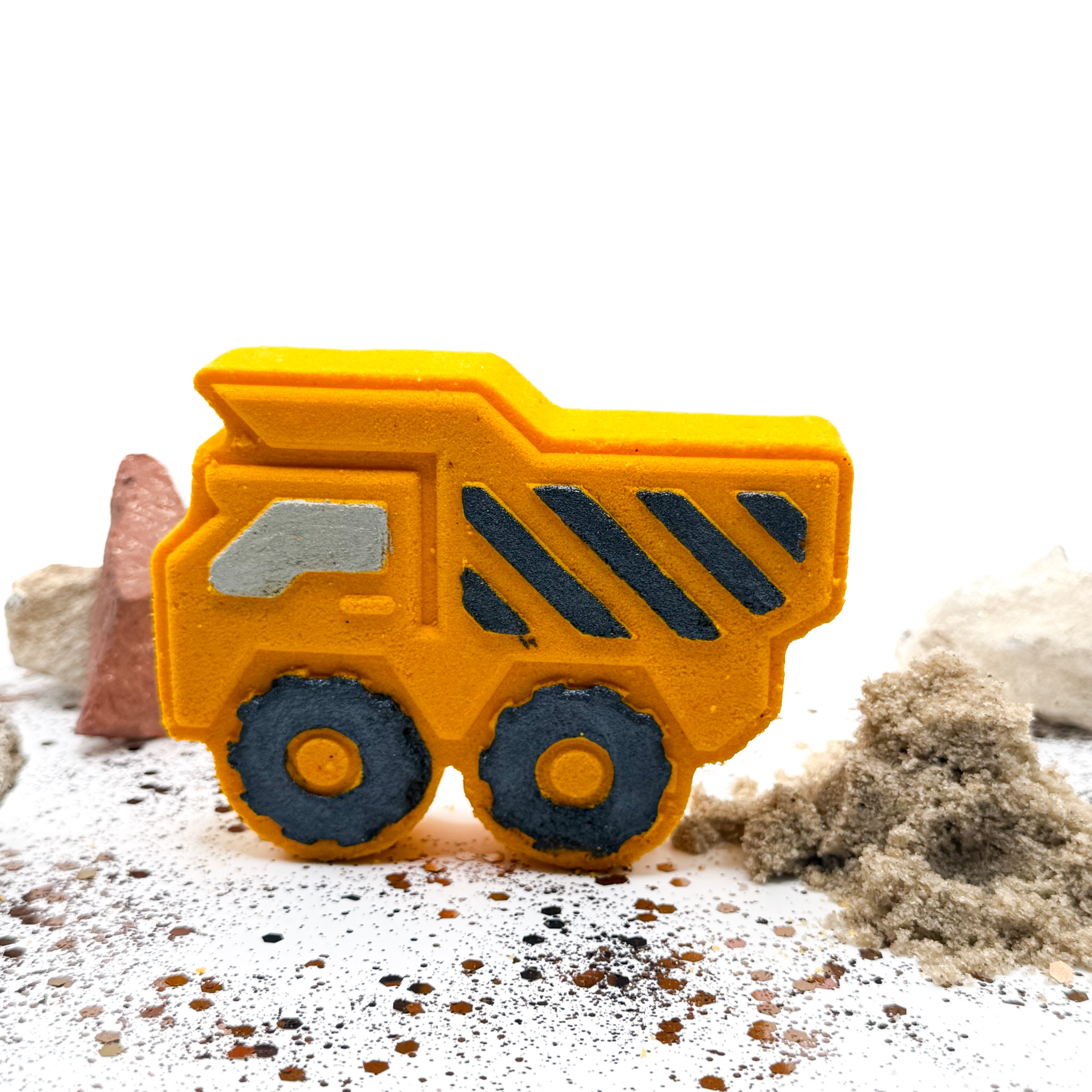 Yellow dump trucks bath bombs made by Rusted Acre soap Co. Black hand painted stripes and other details, standing up with rocks behind it.