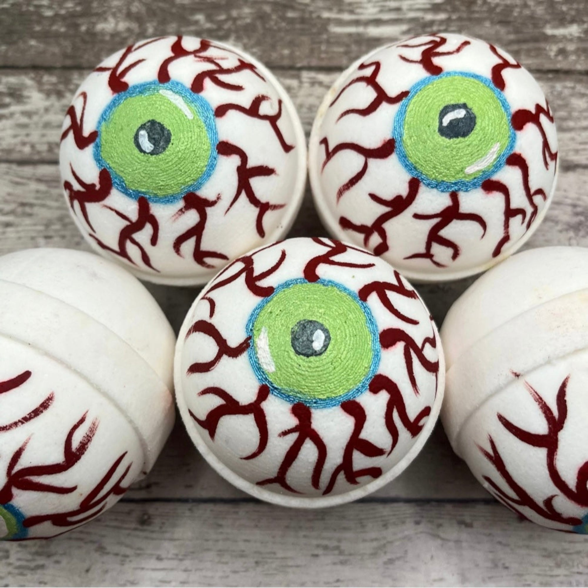 Five hand painted eyeball bath bombs with green irises, blue outlines, and red vein details displayed on a wooden surface made by Rusted Acre Soap Co.